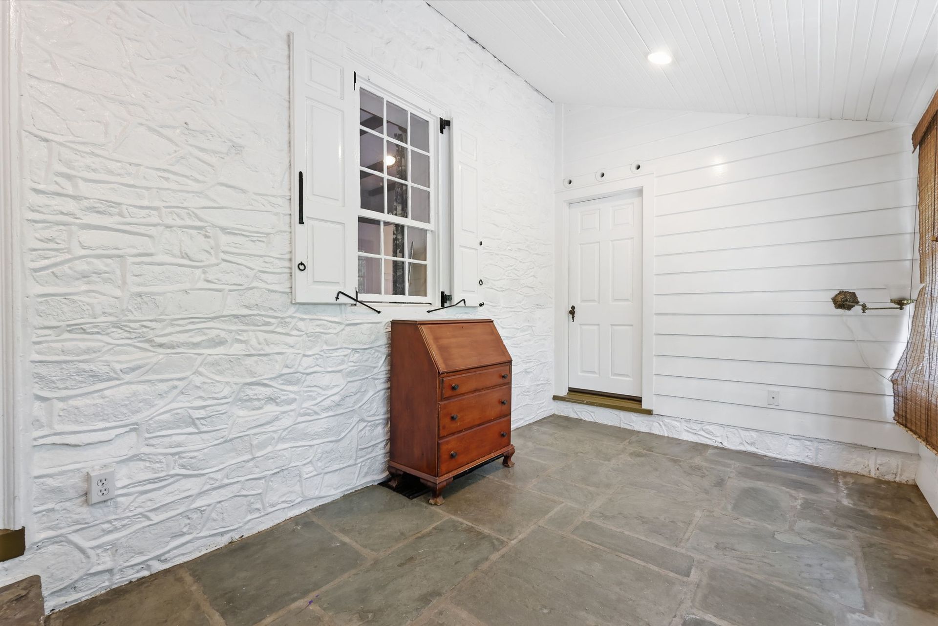 White entry hall with stone floor, white paneled walls, and a small red cabinet beneath a window.