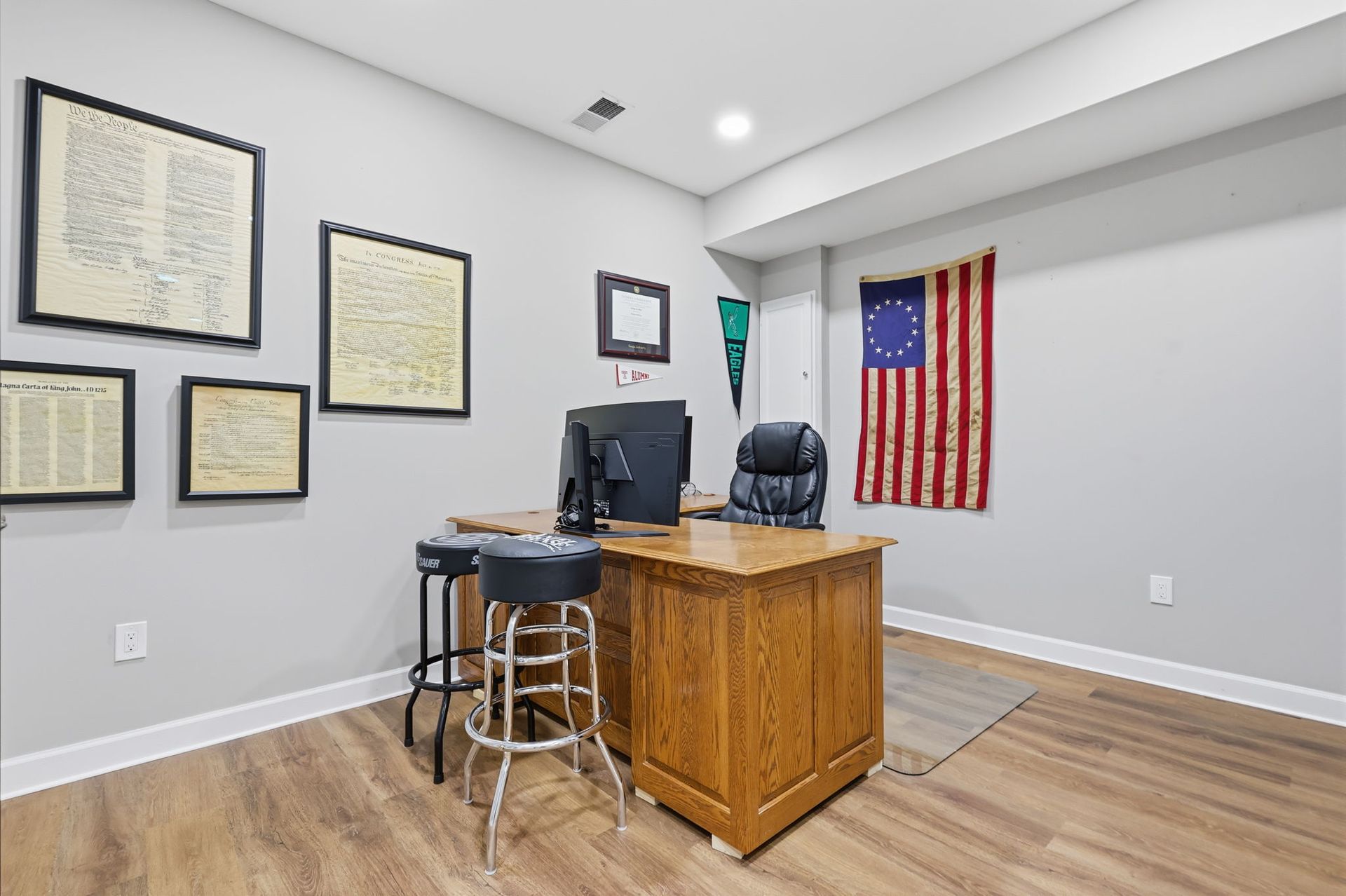 Small office with framed maps, a wooden desk, computer, stool, and an American flag on the wall.