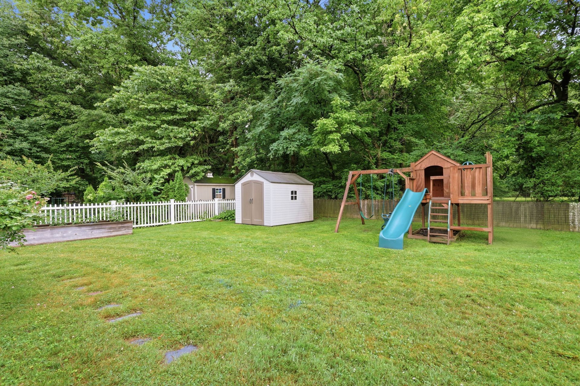 Backyard playground with a blue slide, wooden play structure, shed, and fenced yard under trees