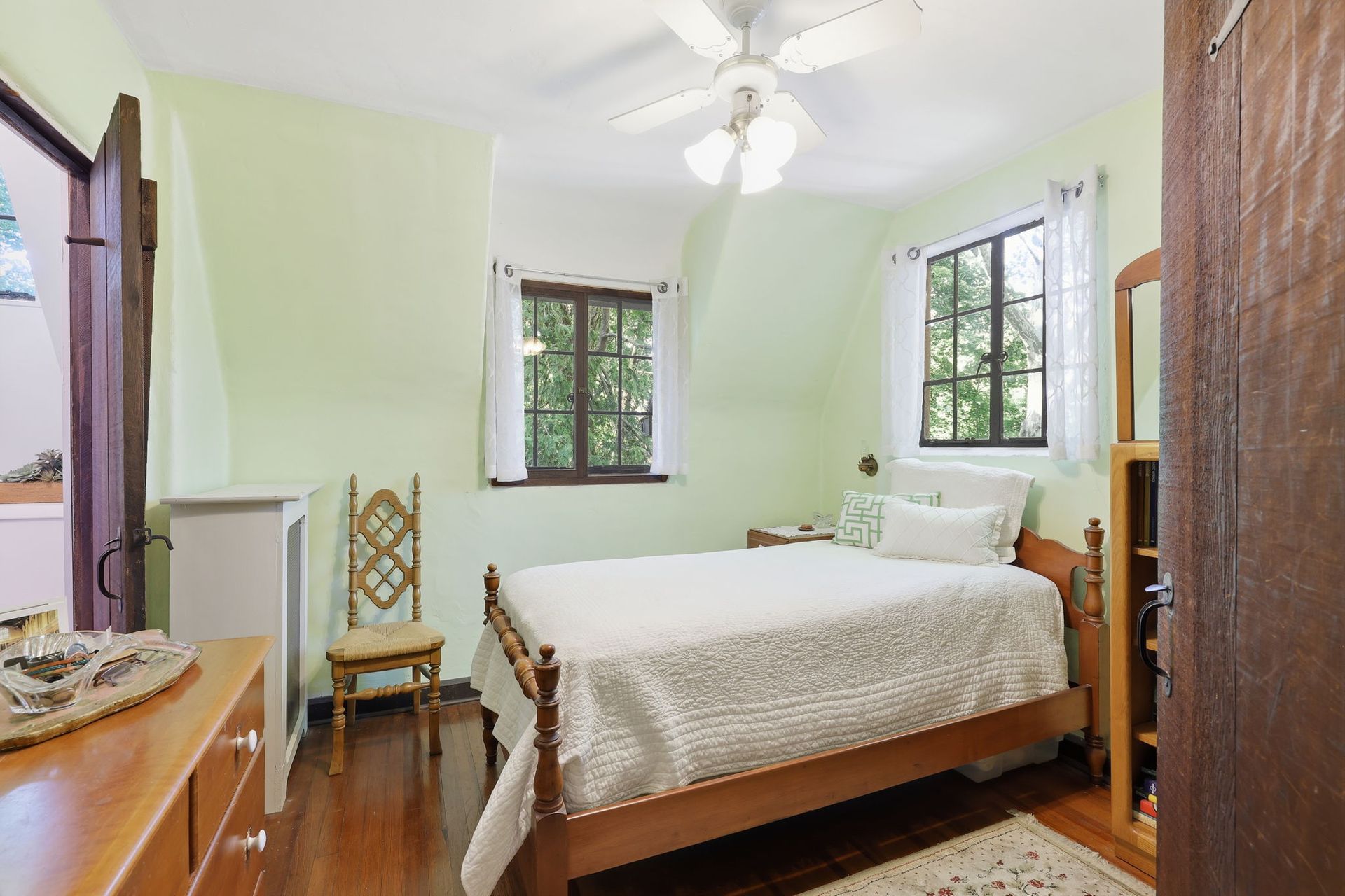 Bright bedroom with white bed, pale green walls, wooden furniture, and open windows.