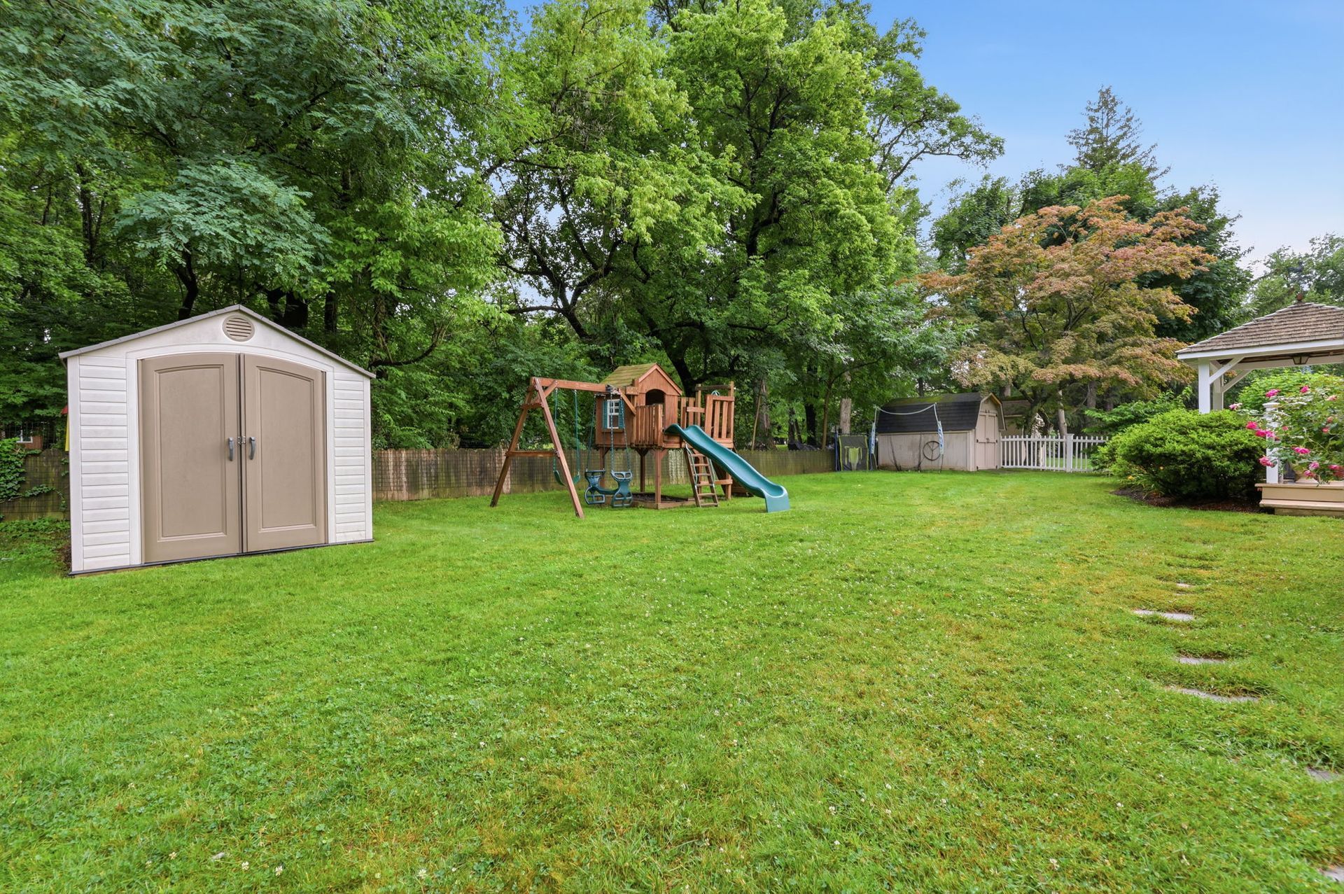 Backyard with a tan storage shed, children’s playset, and large green lawn surrounded by trees.