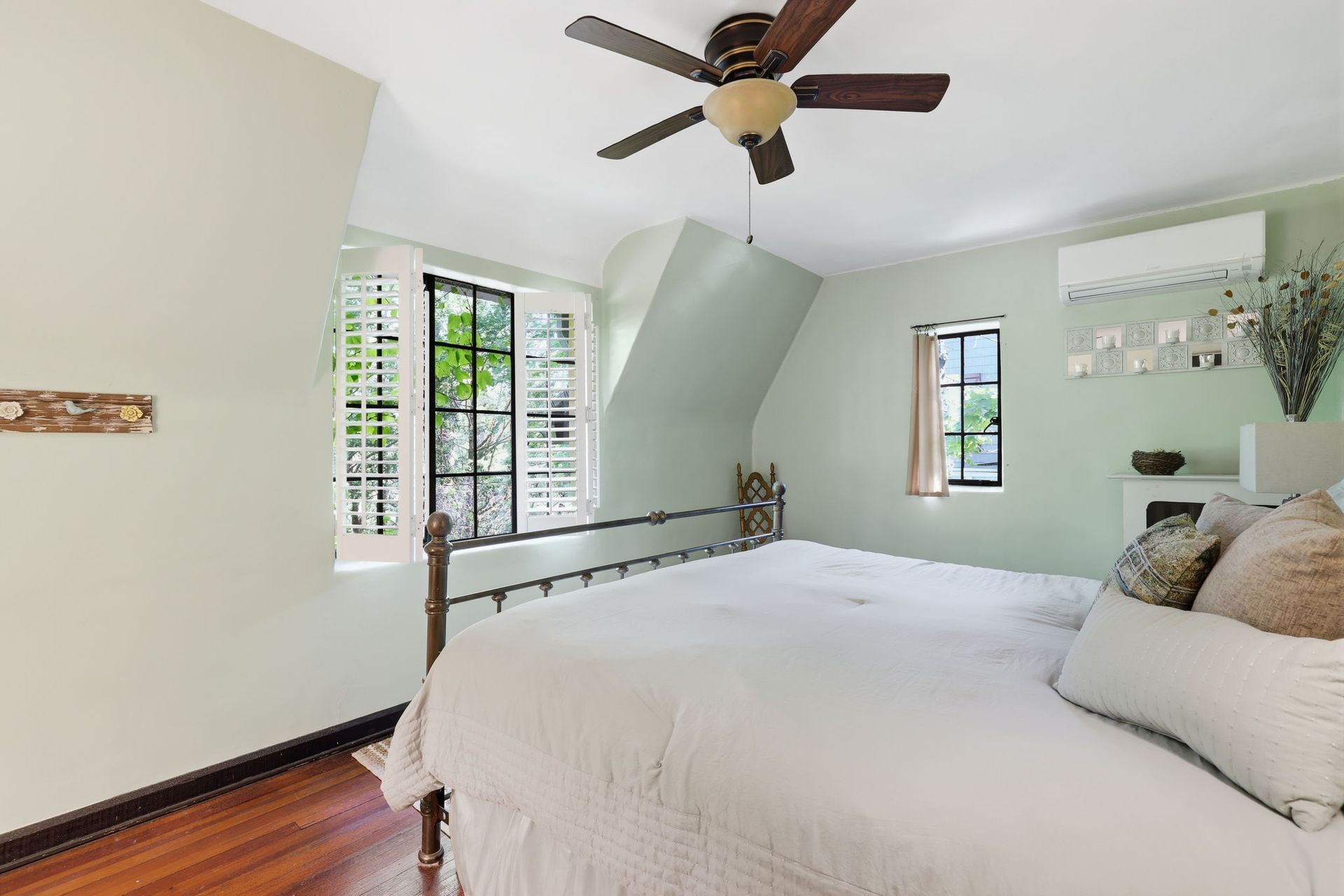 Bright bedroom with white bedding, wood floors, ceiling fan, and windows overlooking greenery