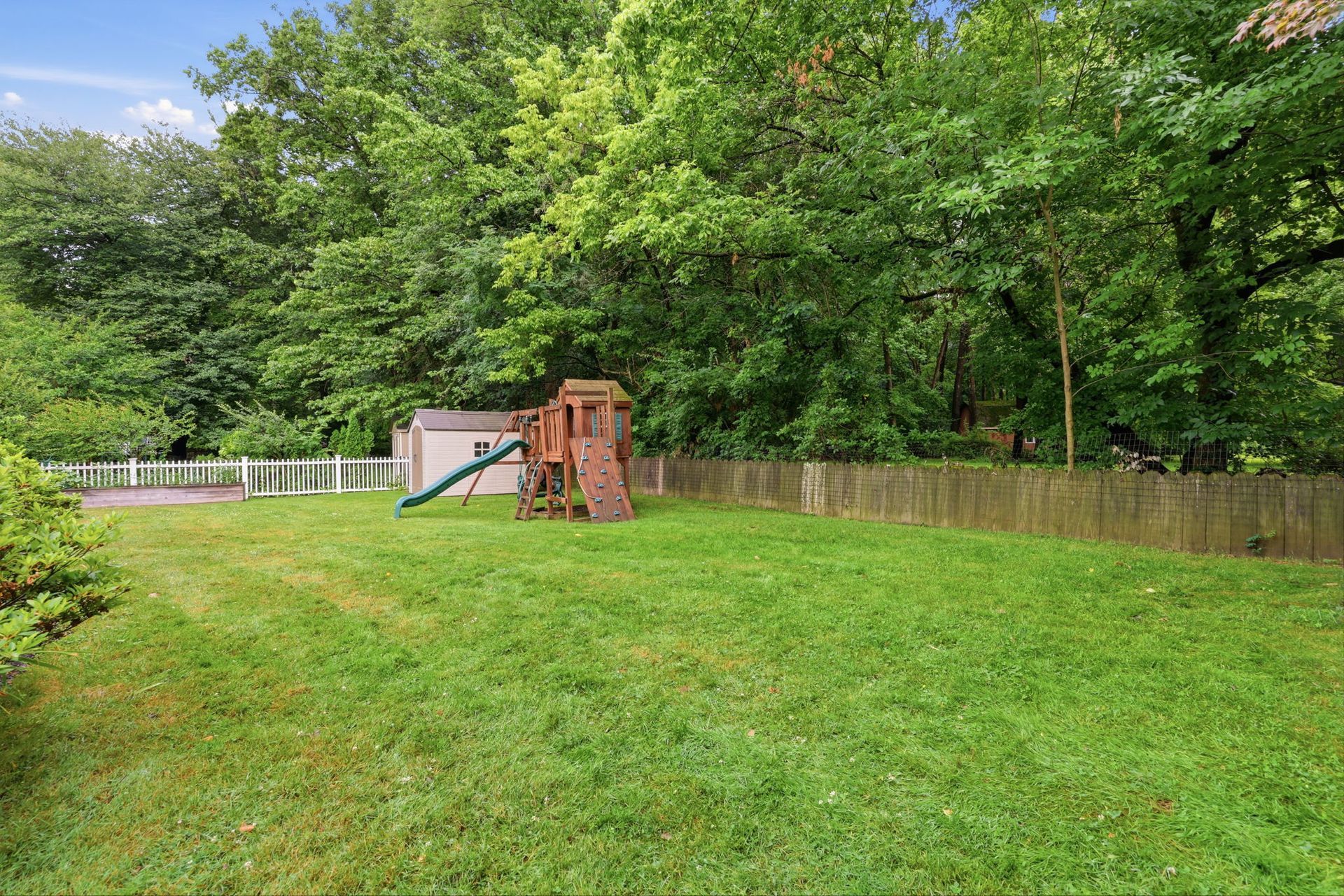 Backyard with green lawn, wooden playset, fence, and trees under a bright sky