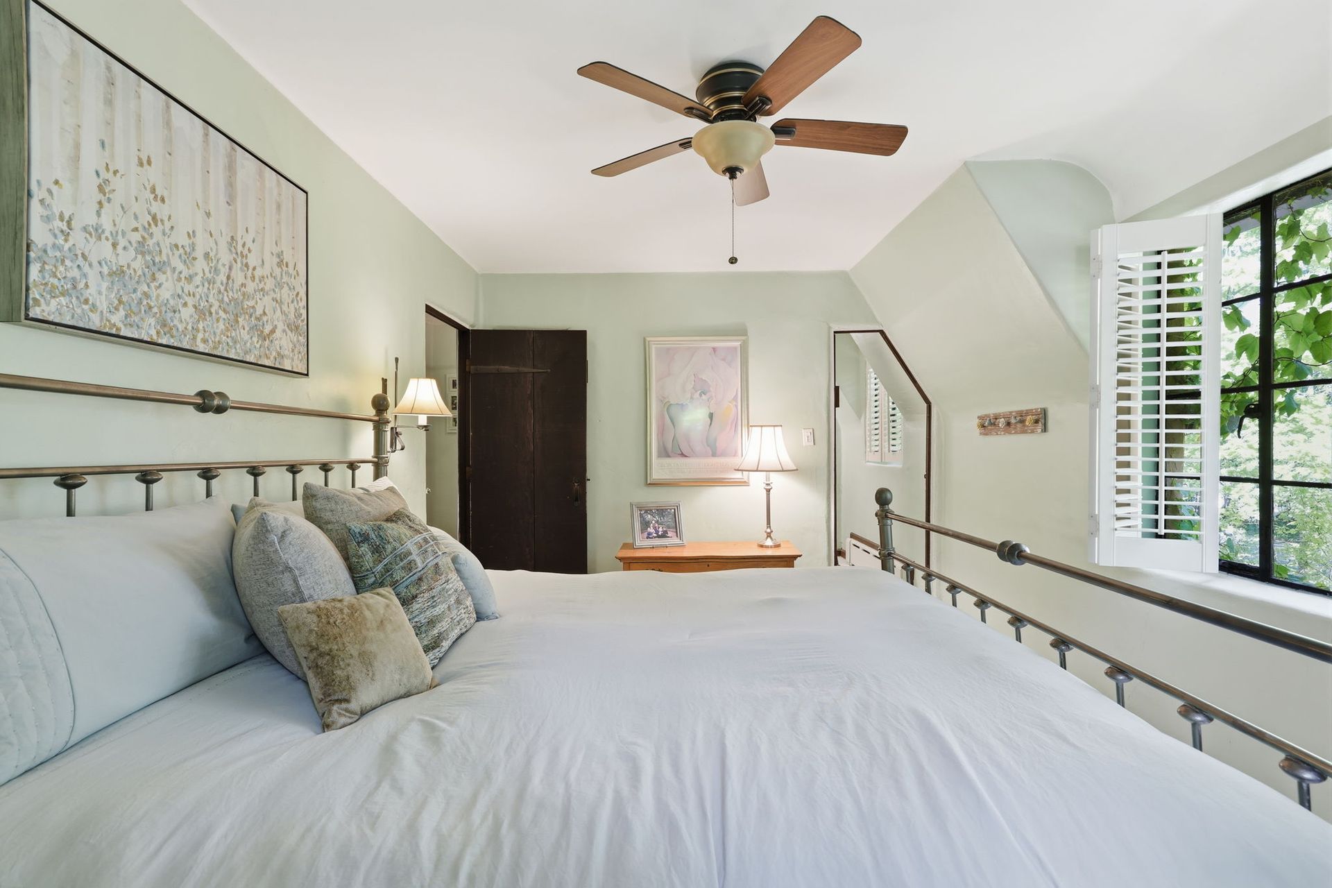 Bright bedroom with white bed, ceiling fan, artwork, and large window with shutters