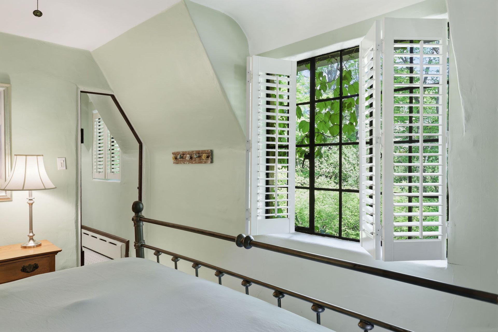 Bright bedroom with a metal bed frame, white shutters, and a window seat by a large bay window.