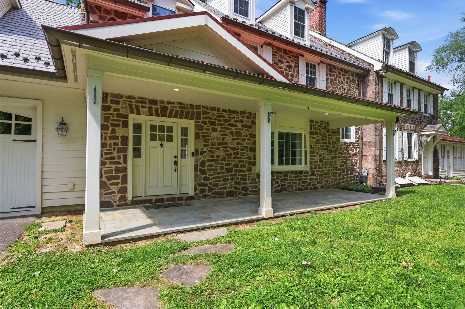 Stone house porch with white columns, green lawn, and red-shingled roof in bright sunlight