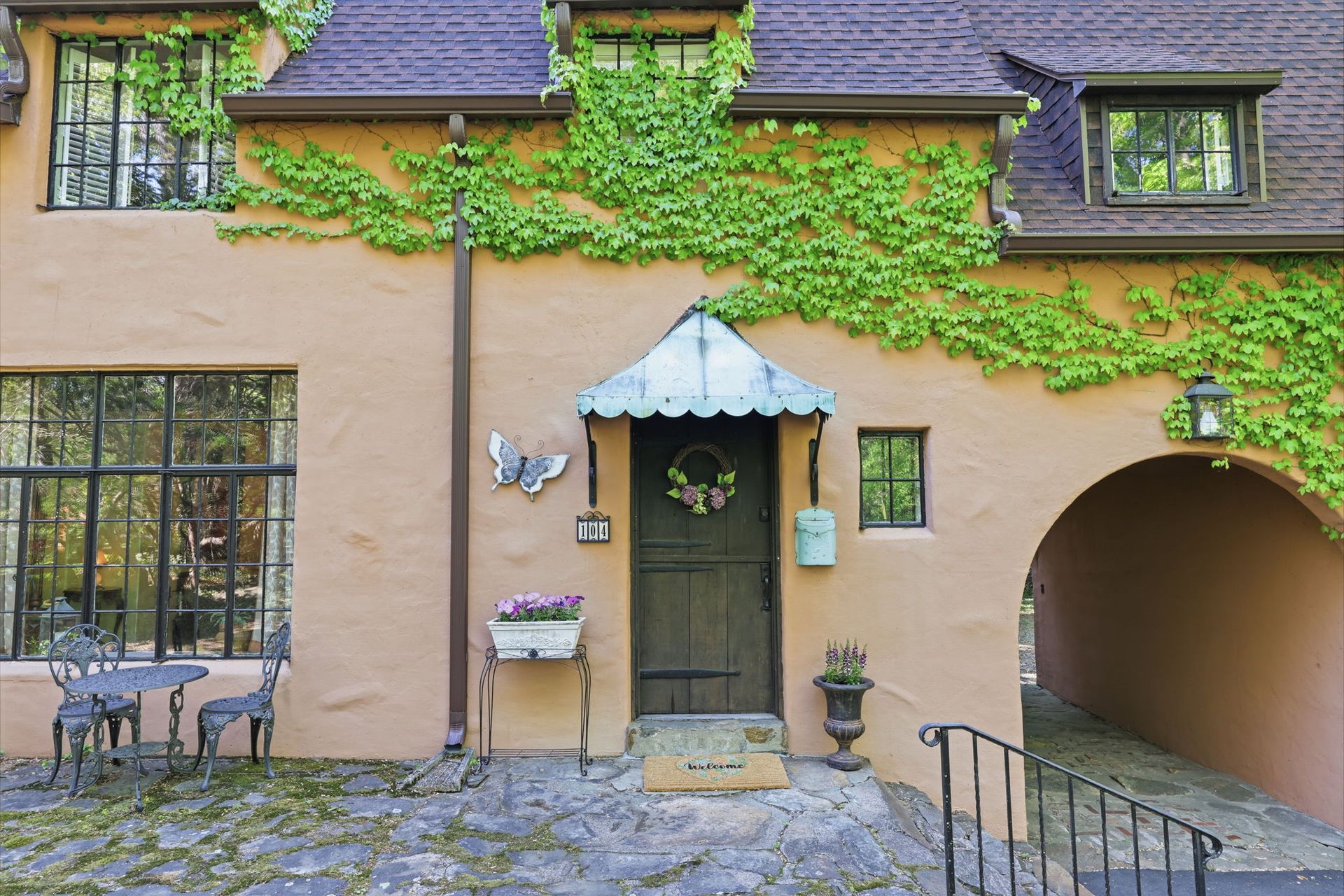 Quaint tan house with ivy, green door, arched passage, and window boxes under a slate roof