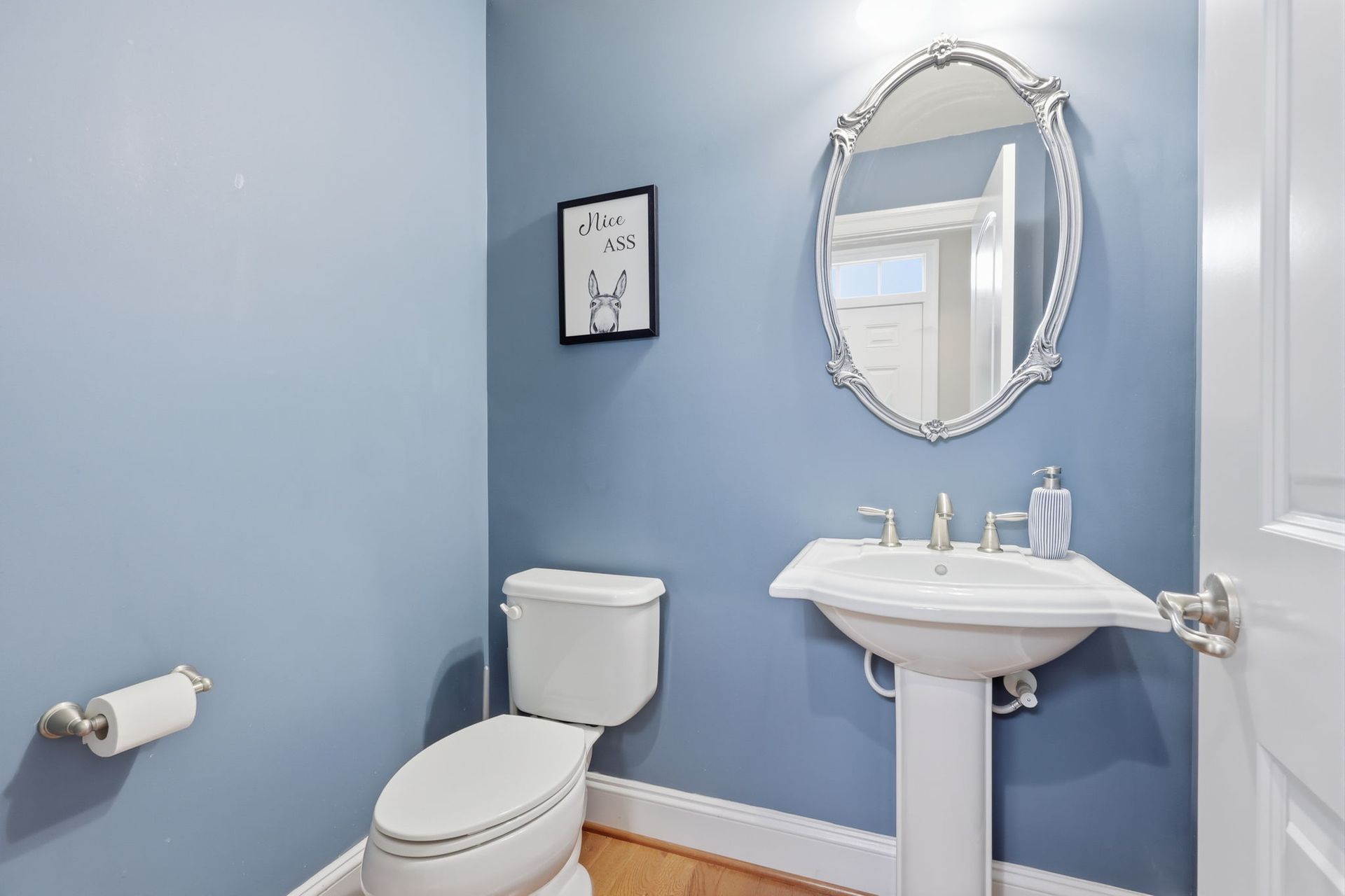 Light blue bathroom with toilet, pedestal sink, and ornate oval mirror.