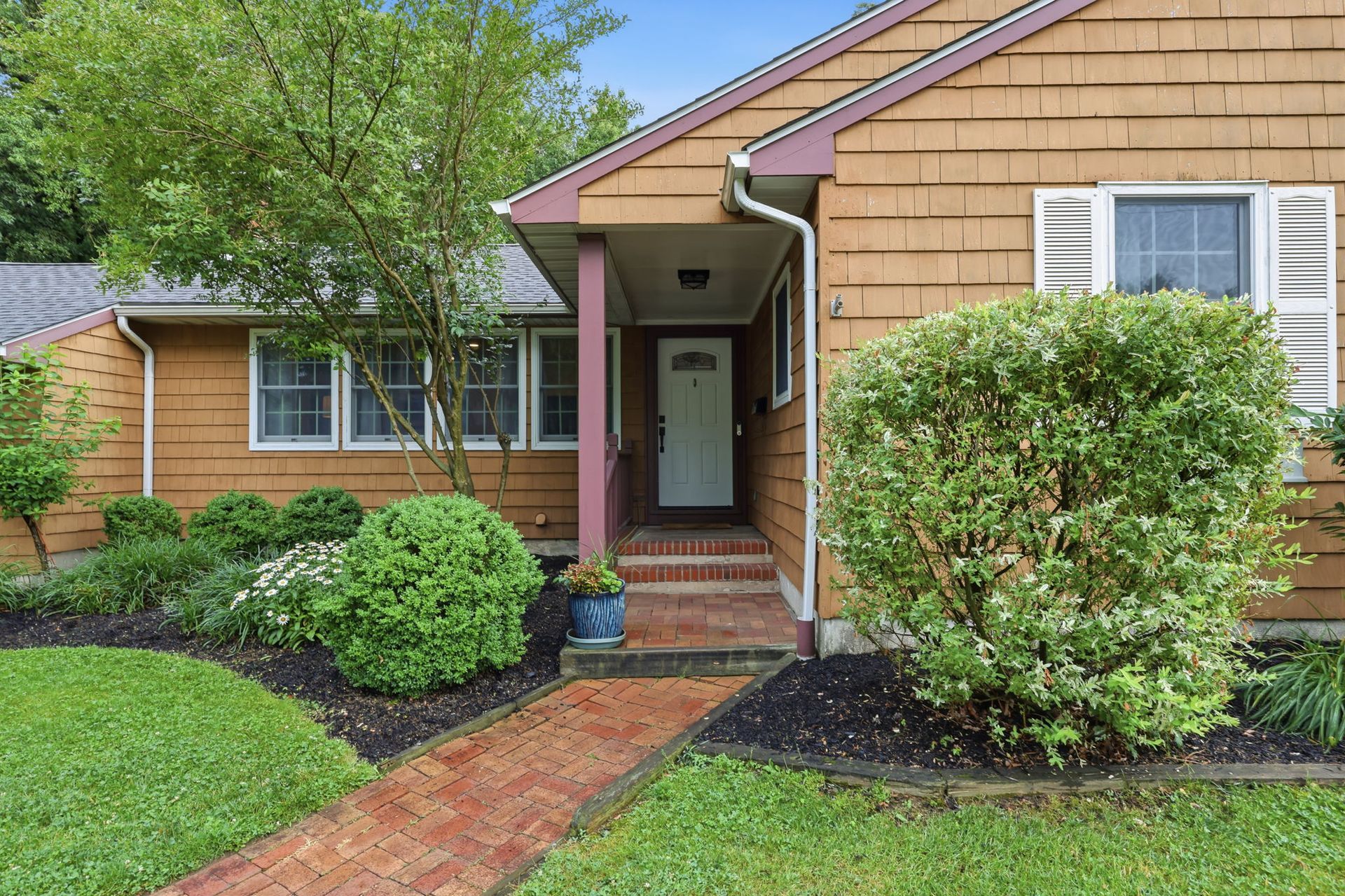 Front entrance of a tan brick house with a red brick walkway and landscaped shrubs