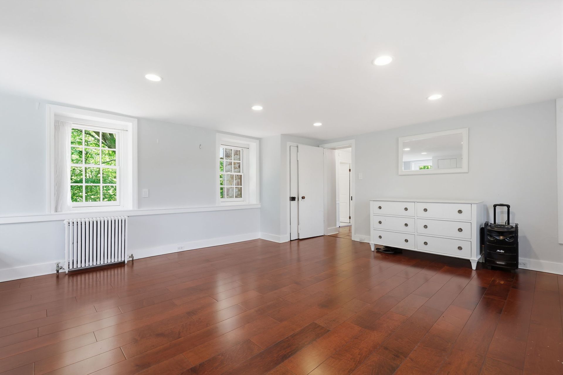Bright empty room with hardwood floors, white walls, windows, and a dresser against the far wall