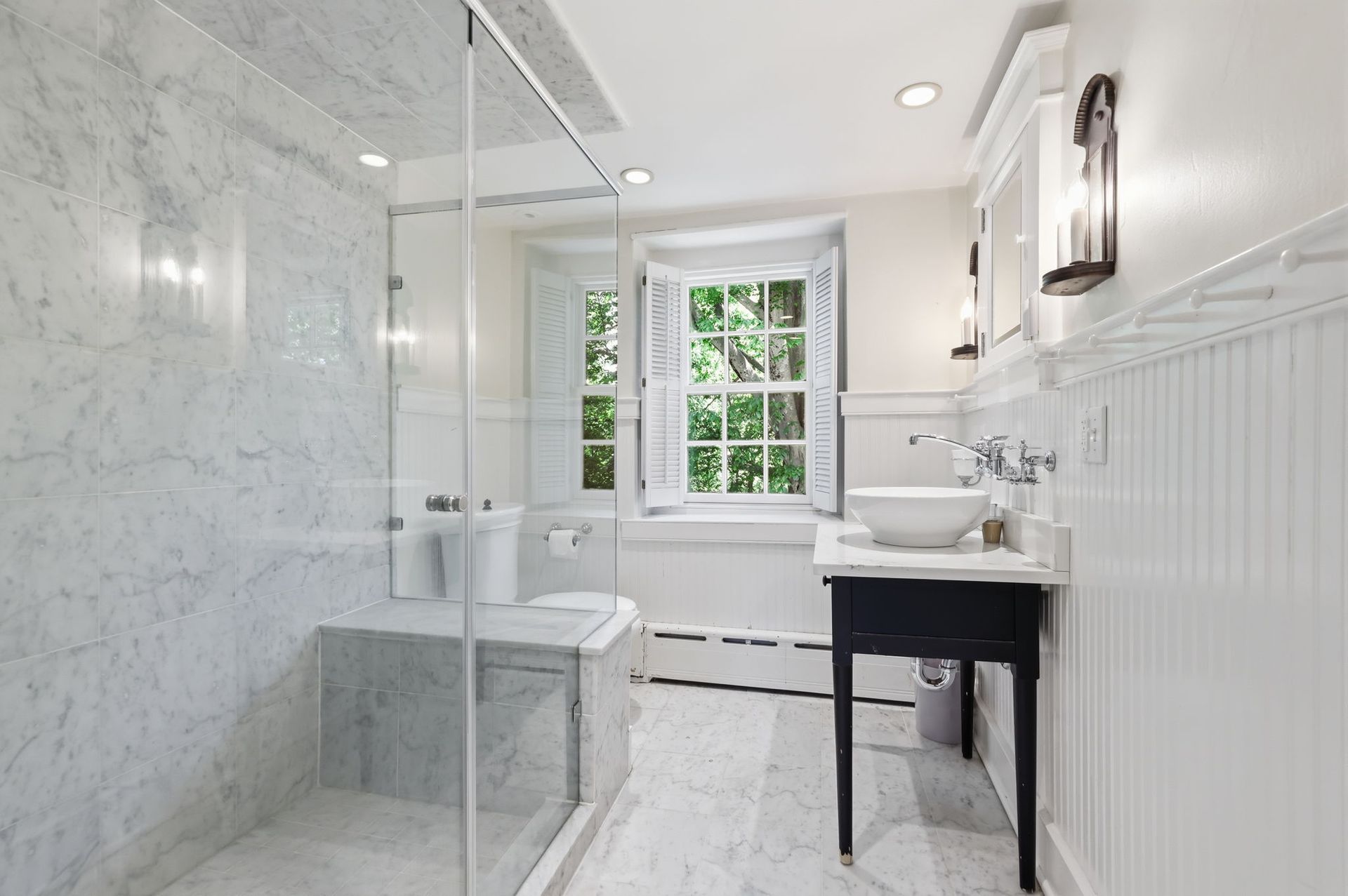 Bright white bathroom with glass shower, marble walls, vanity, and window seat by the window