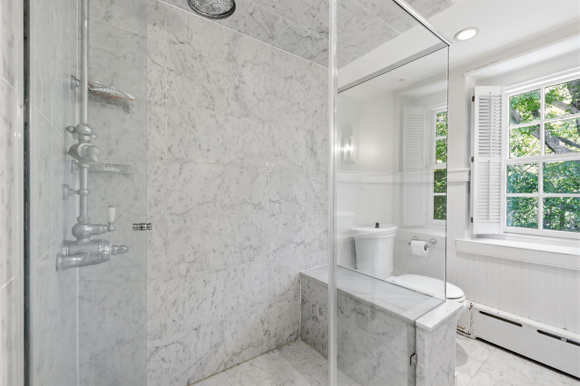 Bright white bathroom with glass shower, toilet, and window letting in daylight.