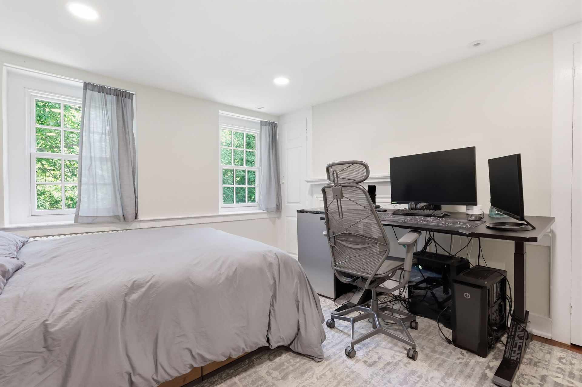 Bright bedroom with bed, desk, dual monitors, and office chair by two windows.