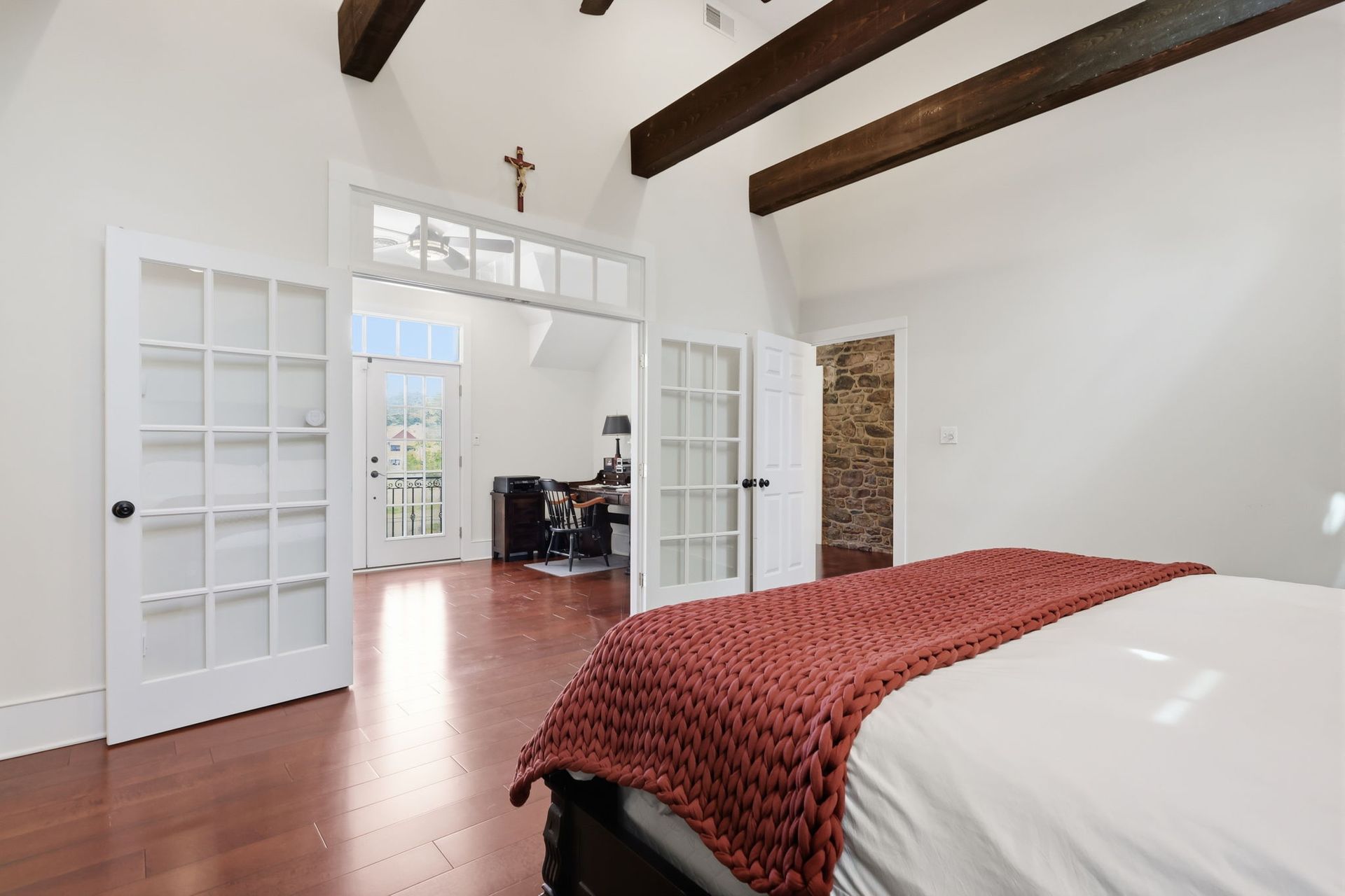 Bright bedroom with white walls, exposed beams, french doors, and a red knit throw on the bed