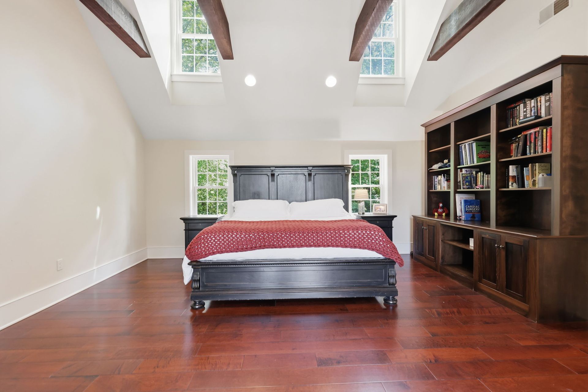 Spacious bedroom with a red bedspread, hardwood floors, high white walls, and a wooden bookshelf cabinet