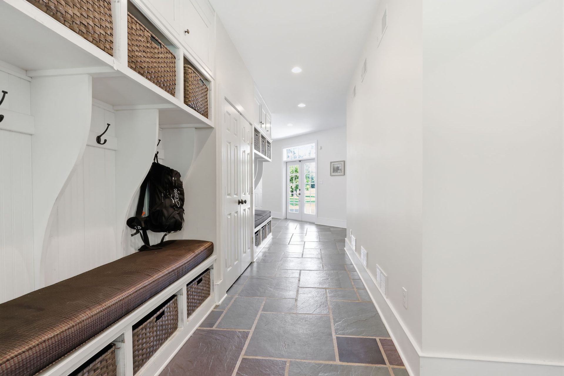 Bright entryway with white walls, slate floor, built-in bench, and coat hooks leading to a sunlit door.