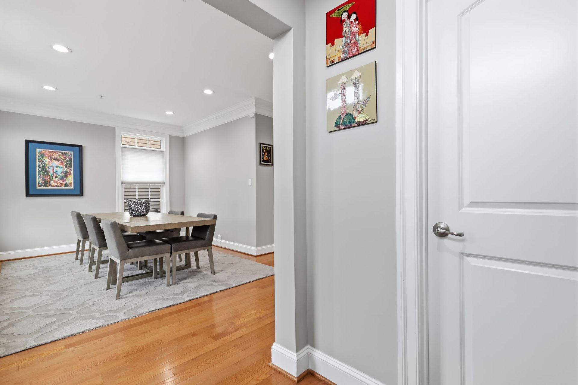 Bright hallway opening to a dining room with wood floors, gray walls, framed art, and a table with chairs.