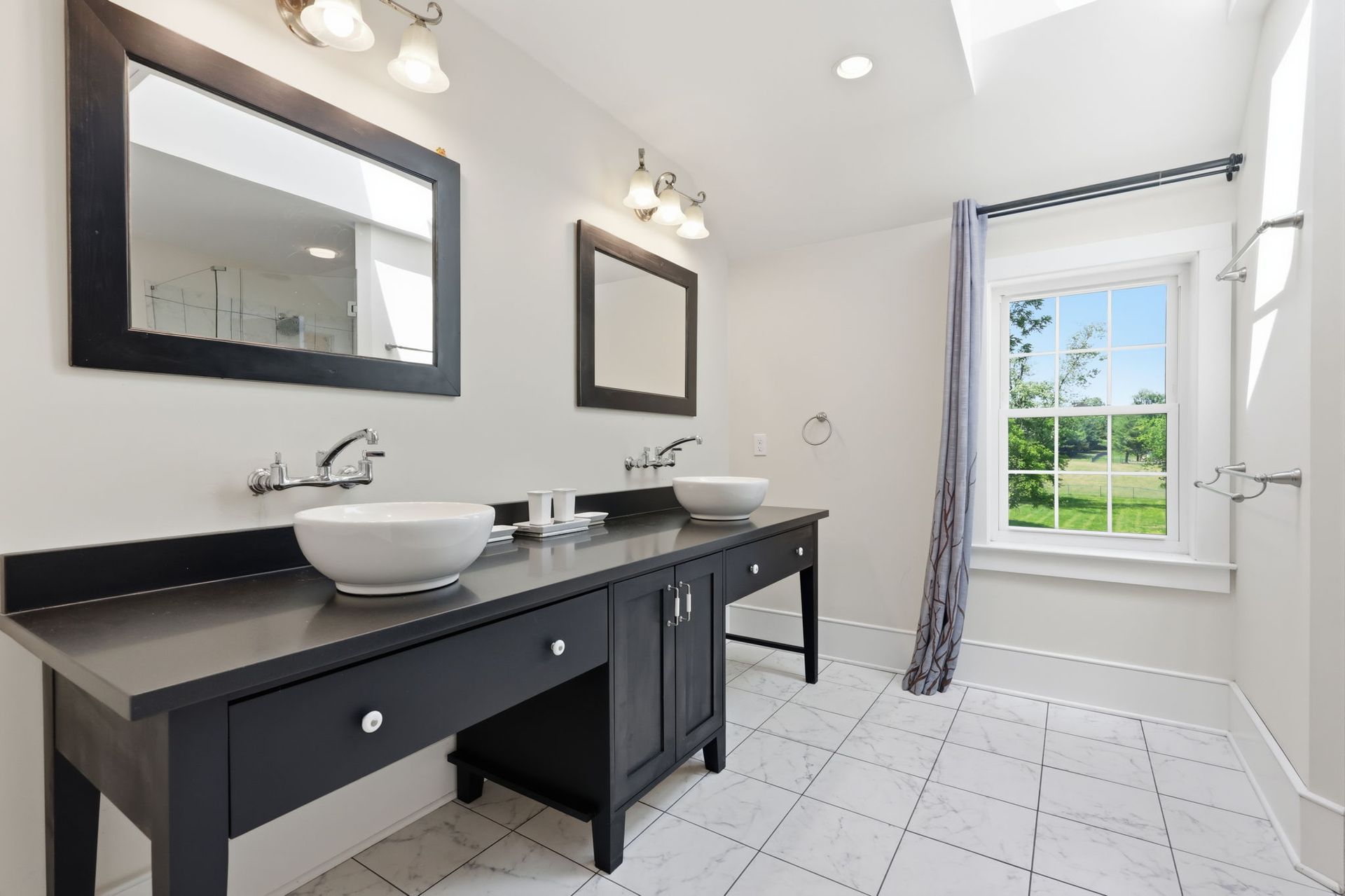 Bright bathroom with double vanity, black countertop, white tile, and window view of green lawn