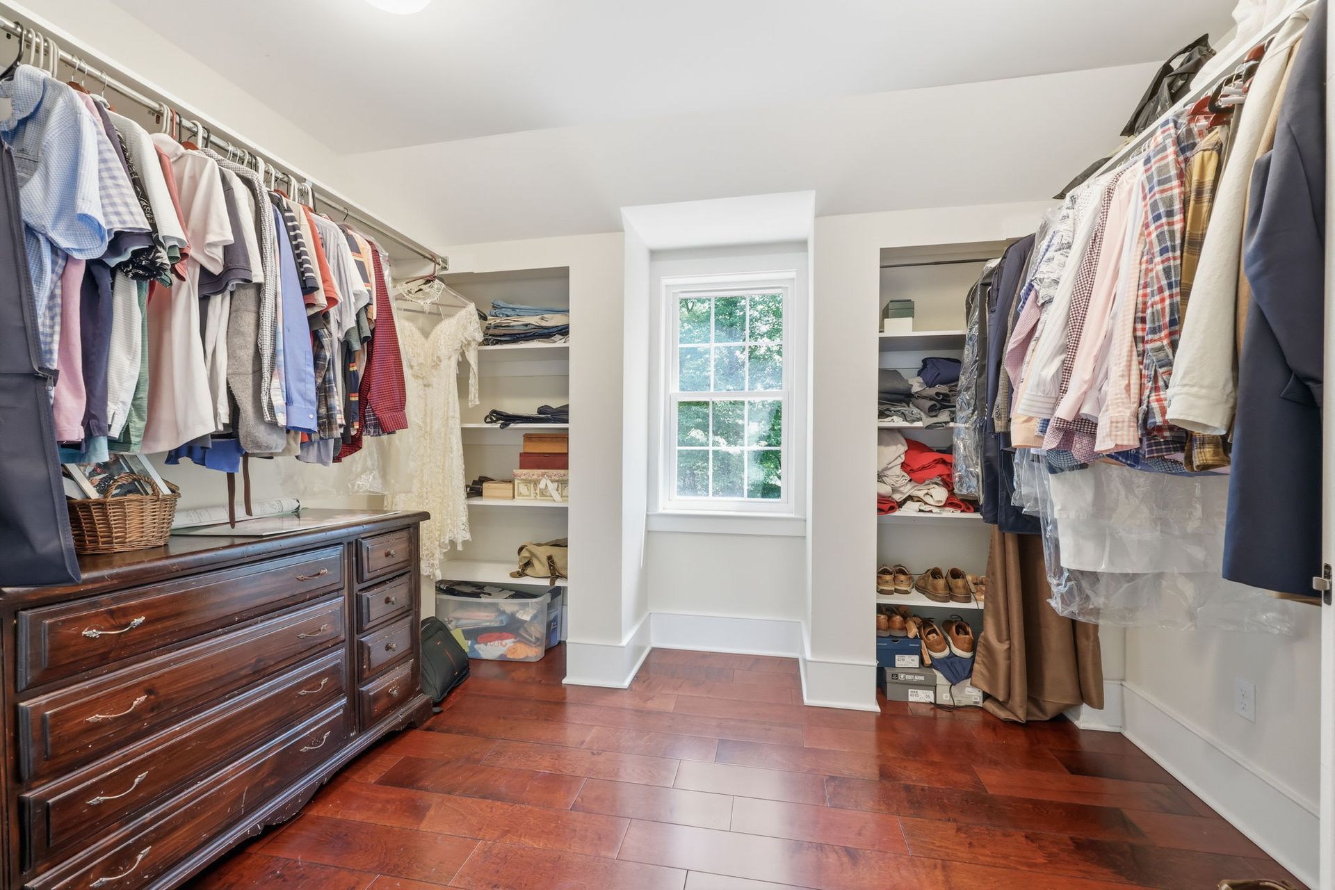 Spacious walk-in closet with hanging clothes, shelves, a wooden dresser, and a centered window with daylight.