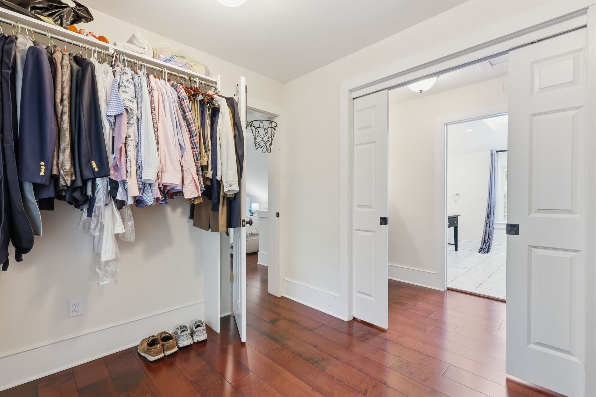 Bright walk-in closet with hanging clothes, shelves, shoes, and open white double doors to a hardwood-floored room