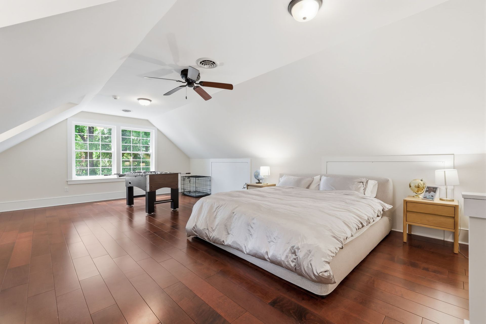 Spacious bedroom with sloped white ceiling, wooden floor, queen bed, desk, and window seat.