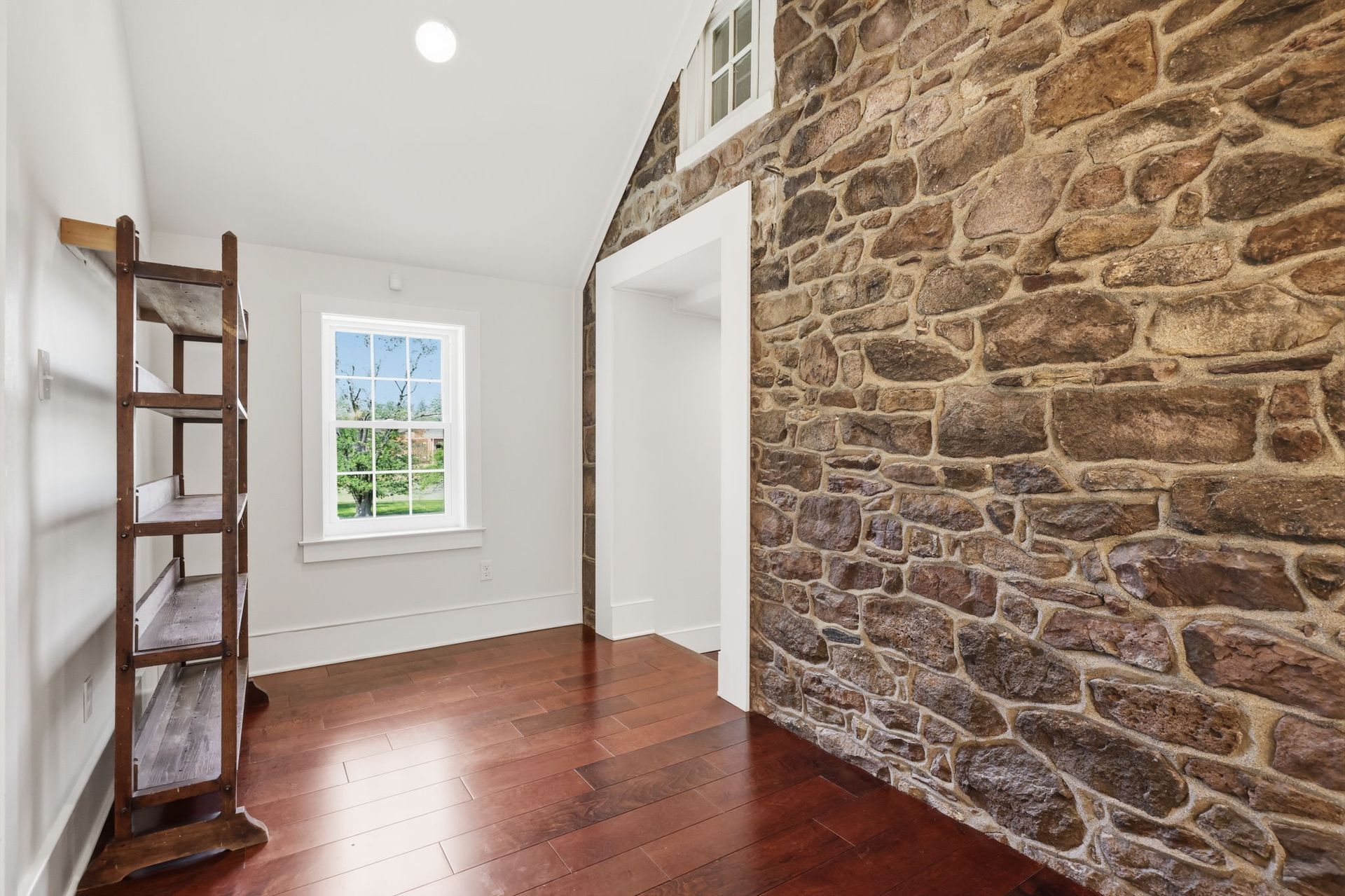 Bright empty room with stone wall, hardwood floor, window, and wooden ladder shelf