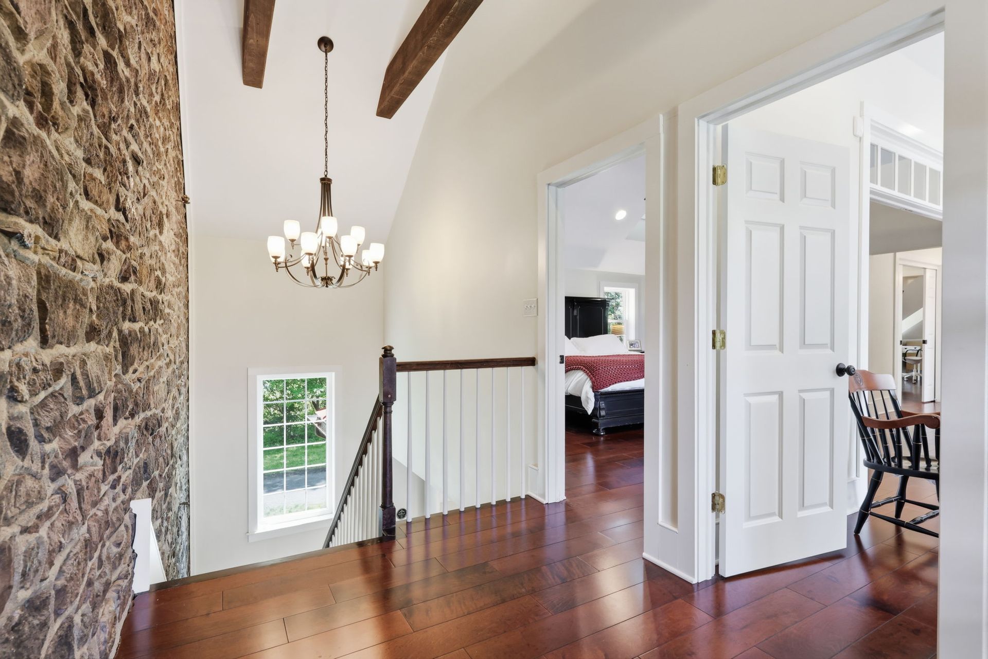 Bright hallway with exposed stone wall, wood floor, chandelier, and open doorway to a bedroom with red bedspread