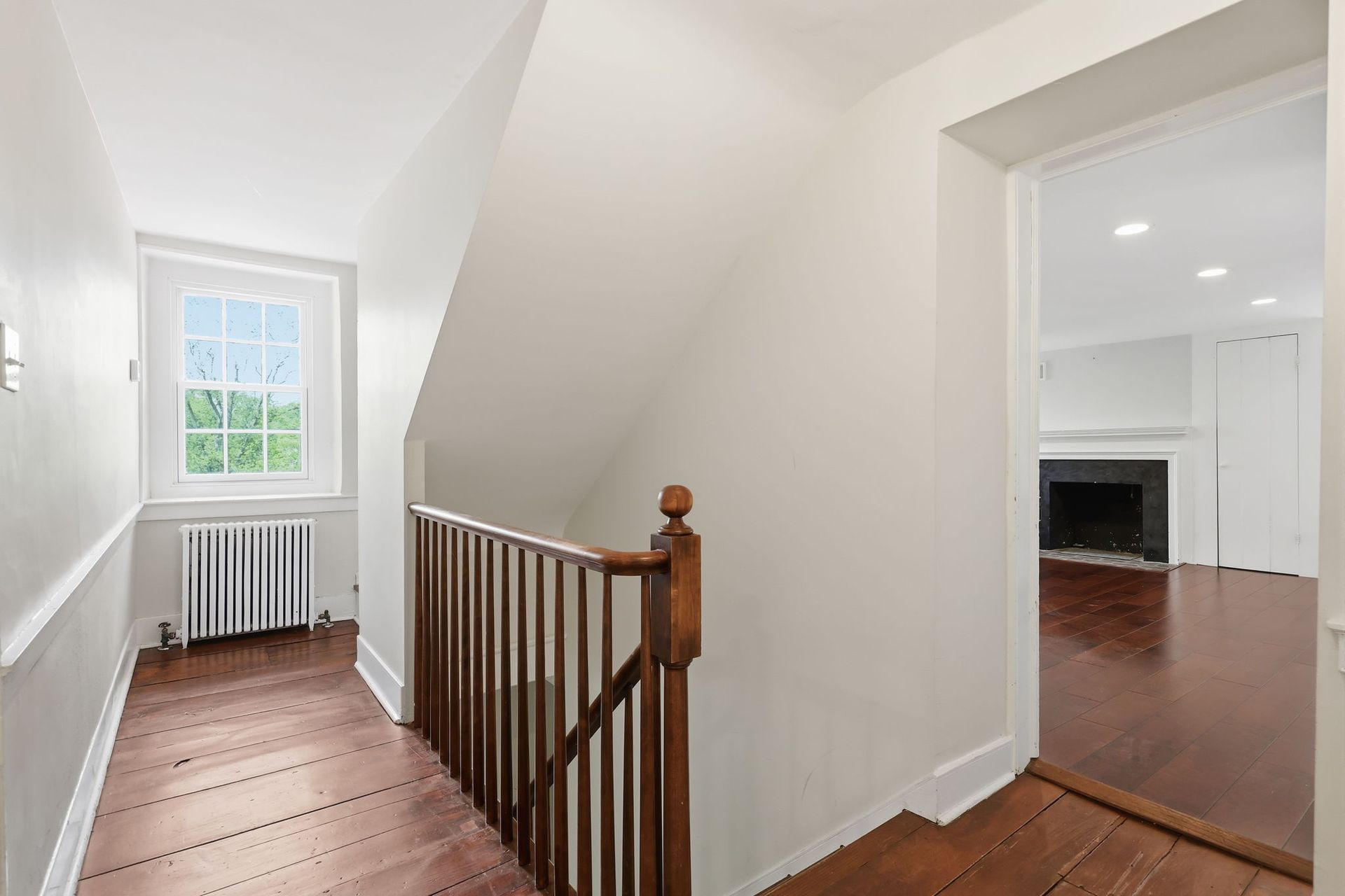 White hallway with wood floor, window and radiator, overlooking a room with a fireplace.