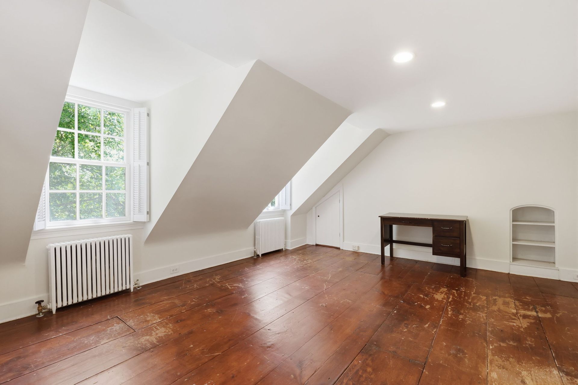 Empty attic room with slanted white walls, wooden floor, a window, and a radiator