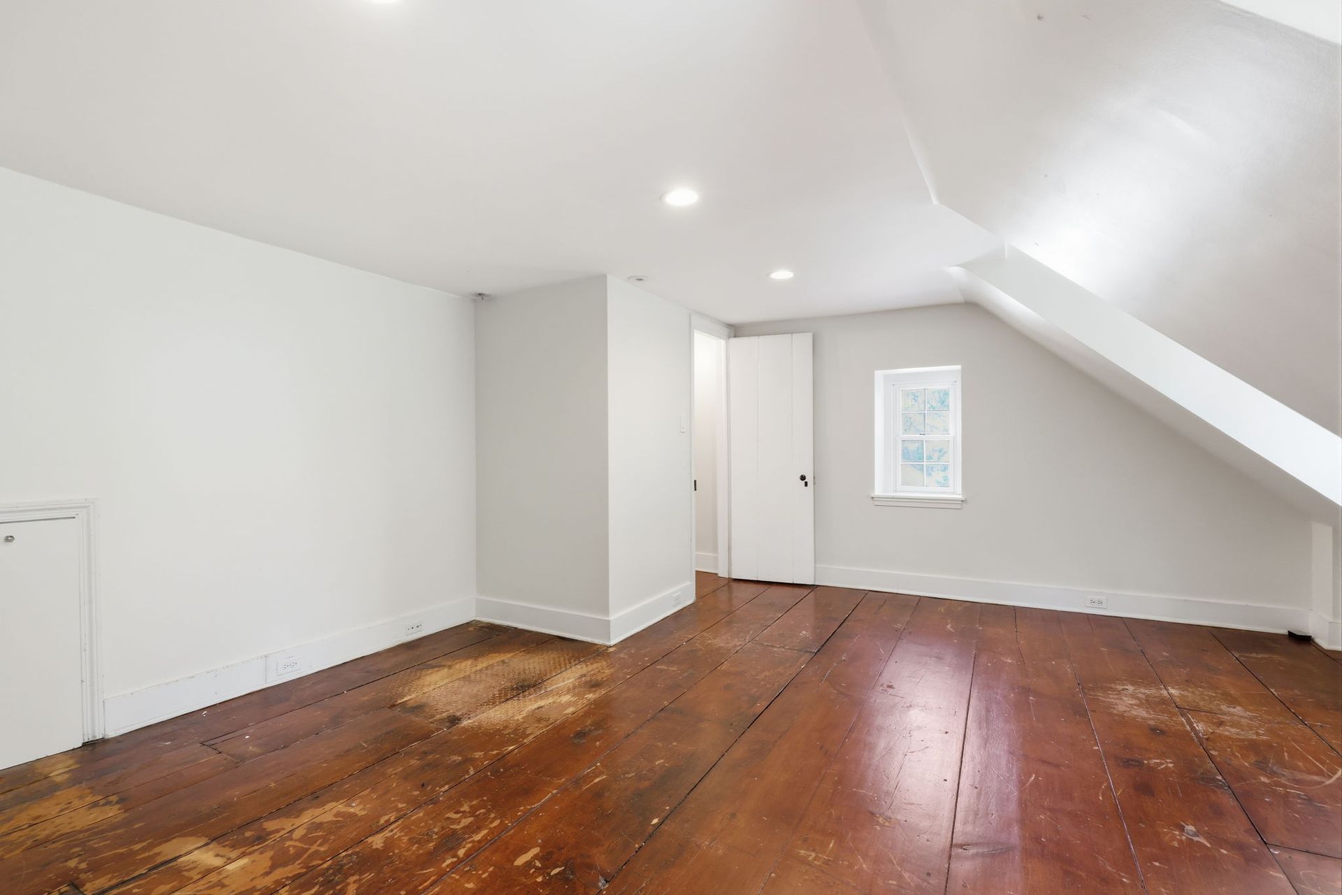 Empty attic room with slanted white ceiling, white walls, and worn wooden floors.
