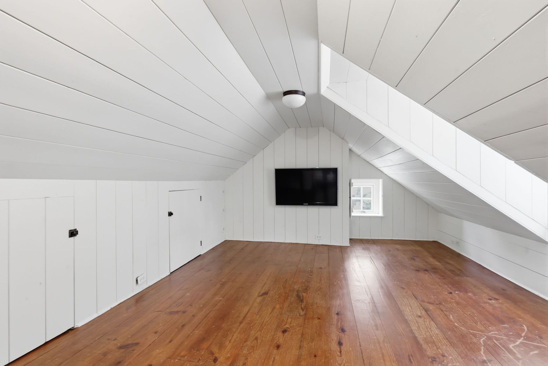 Empty white attic room with sloped ceiling, wood floor, and wall-mounted TV