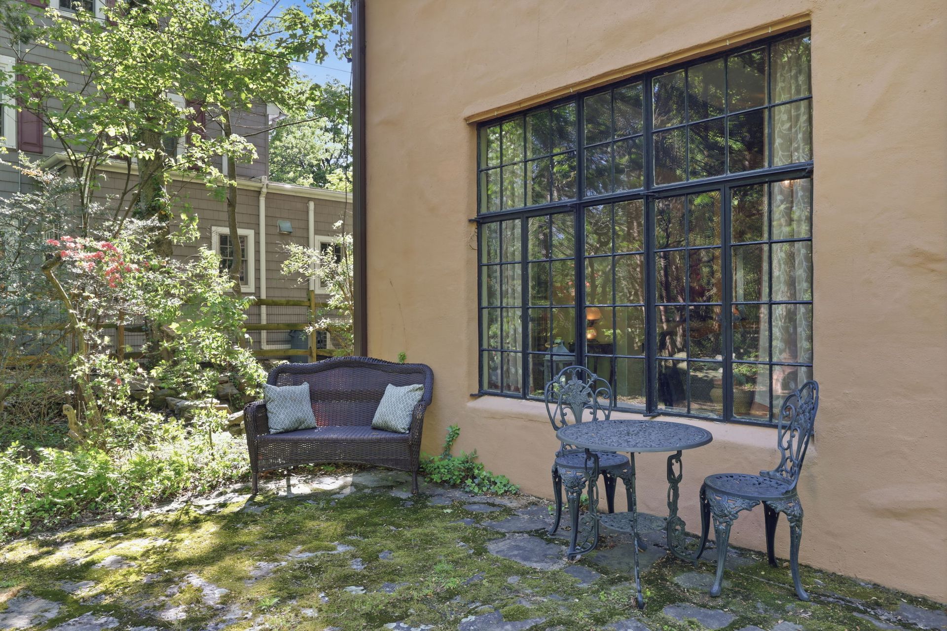 Patio with black metal table and chairs beside a beige house and large window, with a wicker loveseat in the yard