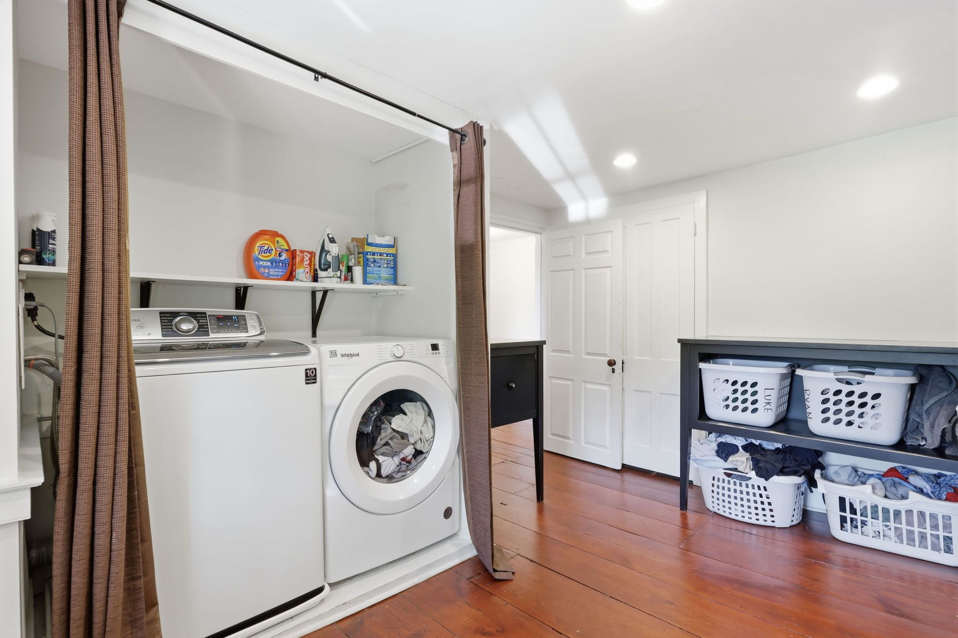 Laundry room with washer, dryer, shelving, and storage baskets on a hardwood floor.