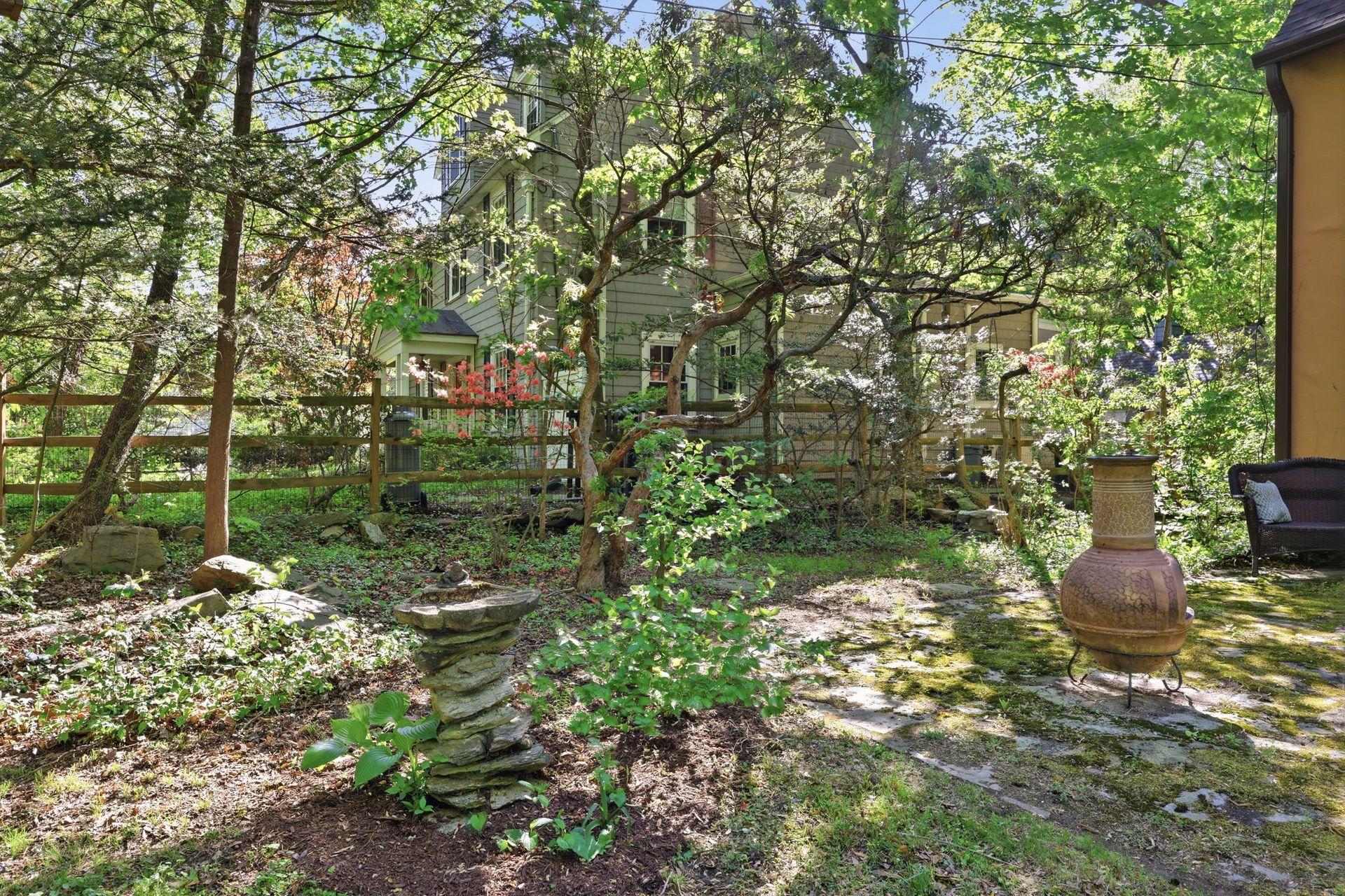 Sunny garden with stone path, trees, fence, and clay pots beside a rustic building