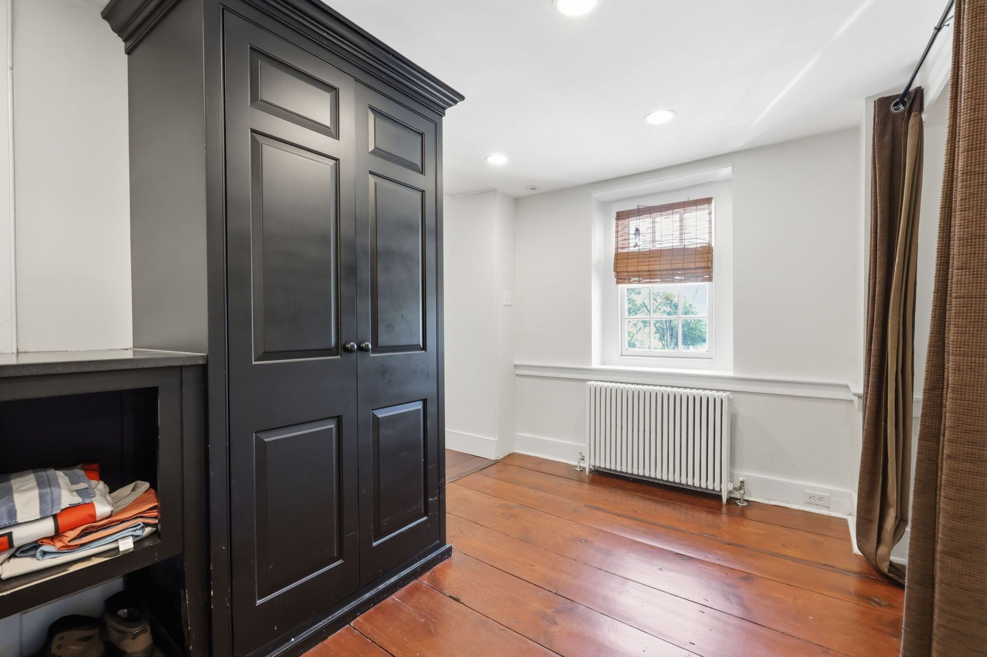 Empty room with black cabinet, white walls, hardwood floor, and a window with blinds and radiator