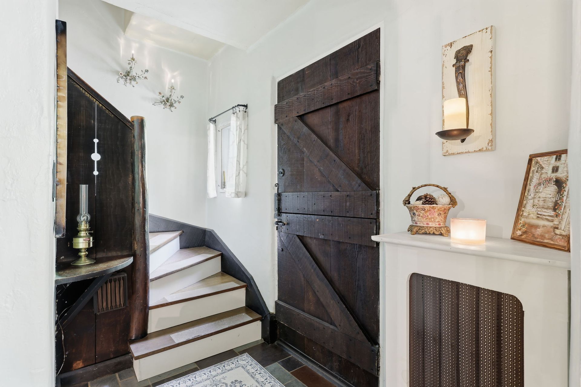 Narrow entryway with stairs, dark wooden door, white walls, and decorative shelves and mirror