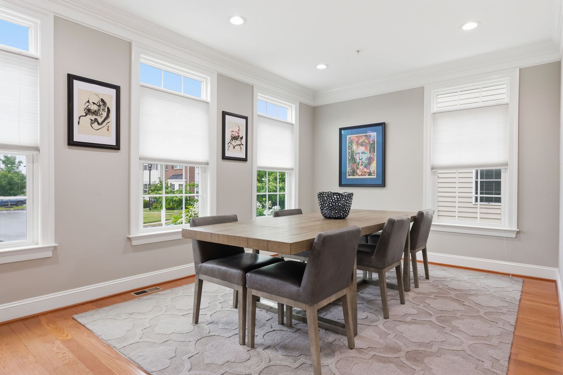 Bright dining room with a wooden table, dark chairs, gray walls, framed art, and large windows