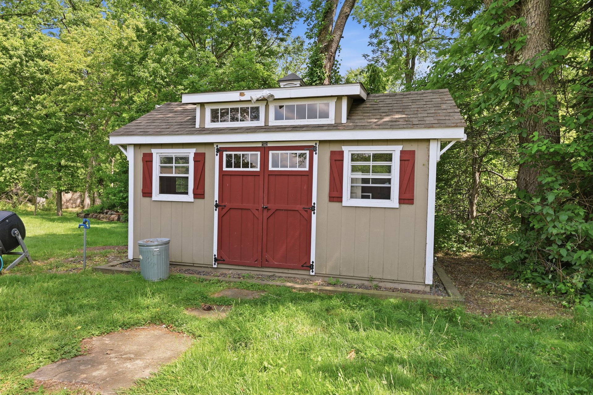 Small red and beige shed with white trim, double doors, and grassy yard surrounded by trees.