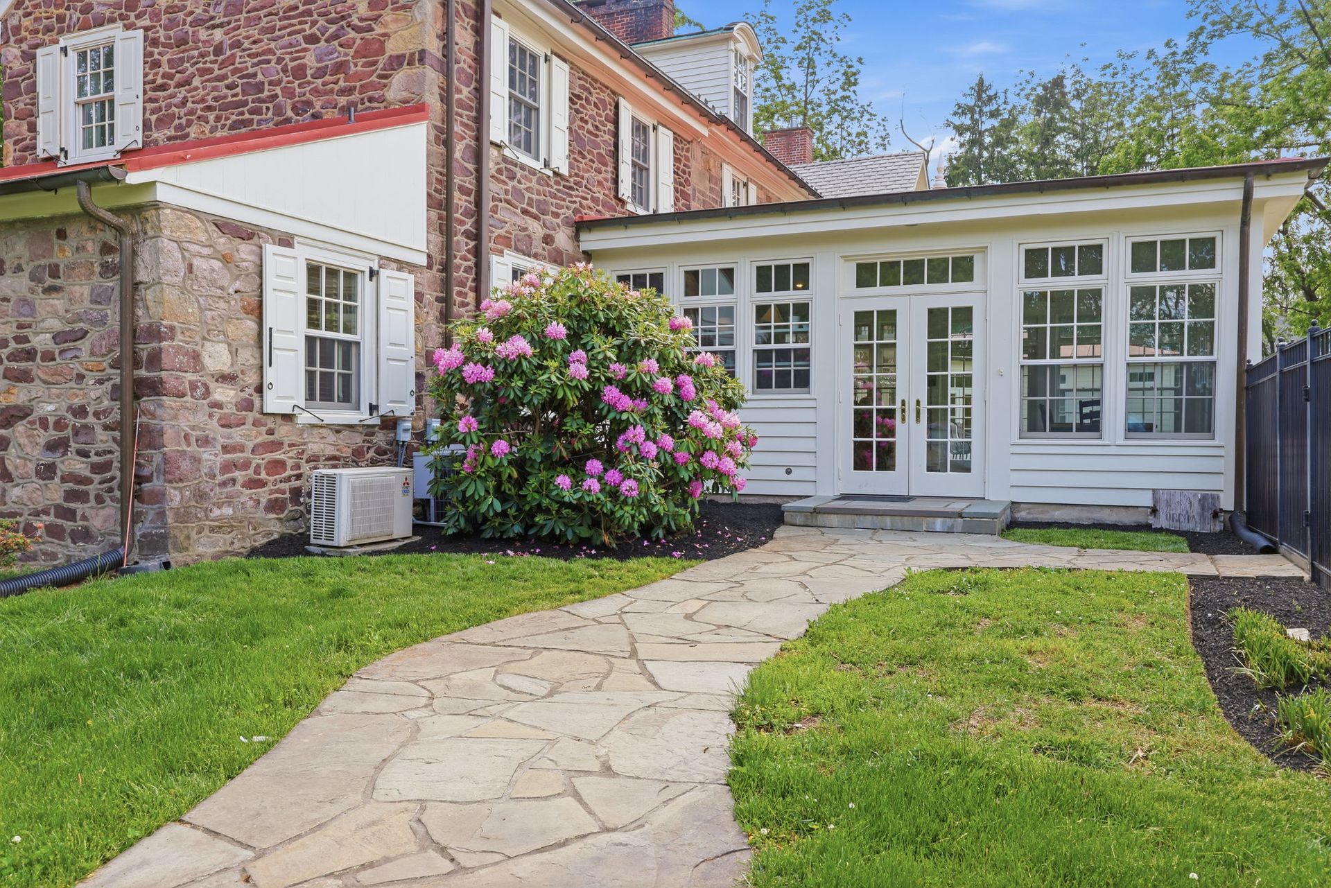 Stone walkway leads to a white sunroom beside a brick house and flowering shrub in a green yard