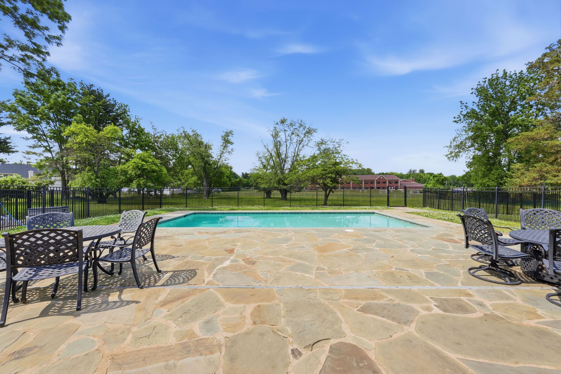 Patio overlooking a fenced swimming pool with stone paving, tables, chairs, and trees under a blue sky