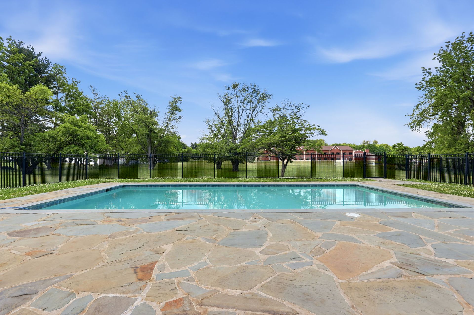 Stone patio beside a turquoise pool with trees and a red-roofed pavilion under a blue sky