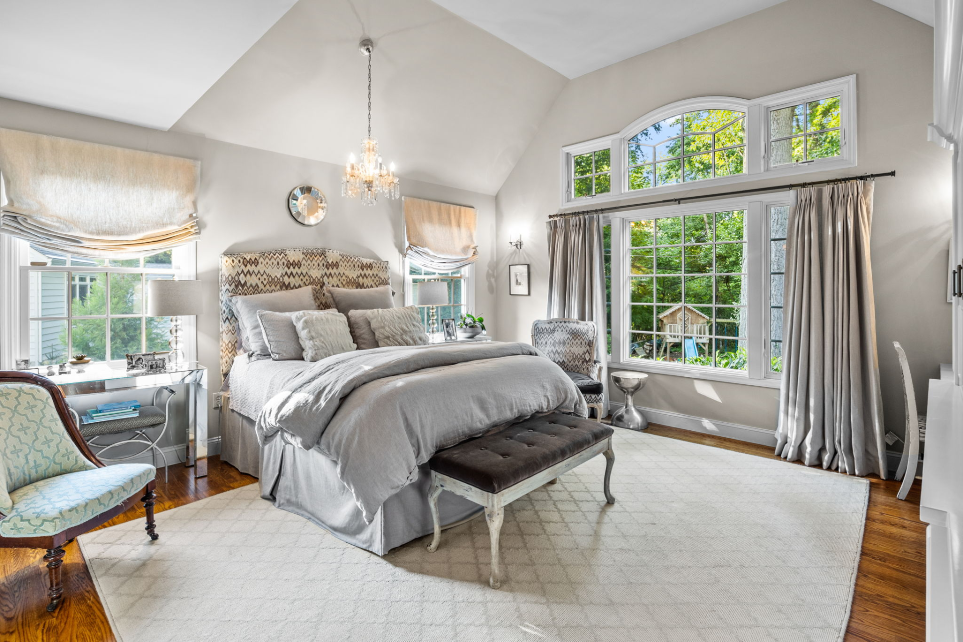 Spacious, light-filled bedroom with bed, windows, chandelier, and seating area. Soft gray and white color scheme.