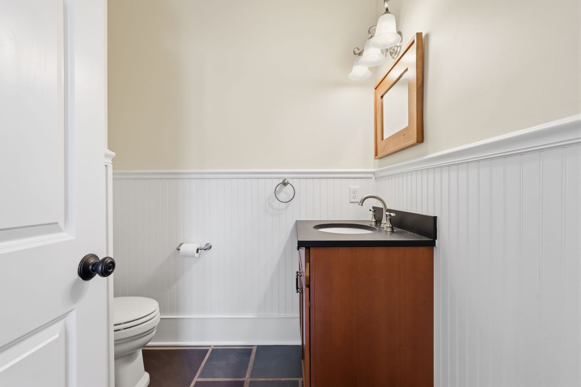 Small bathroom with white wainscoting, dark vanity, sink, toilet, and mirror on the wall