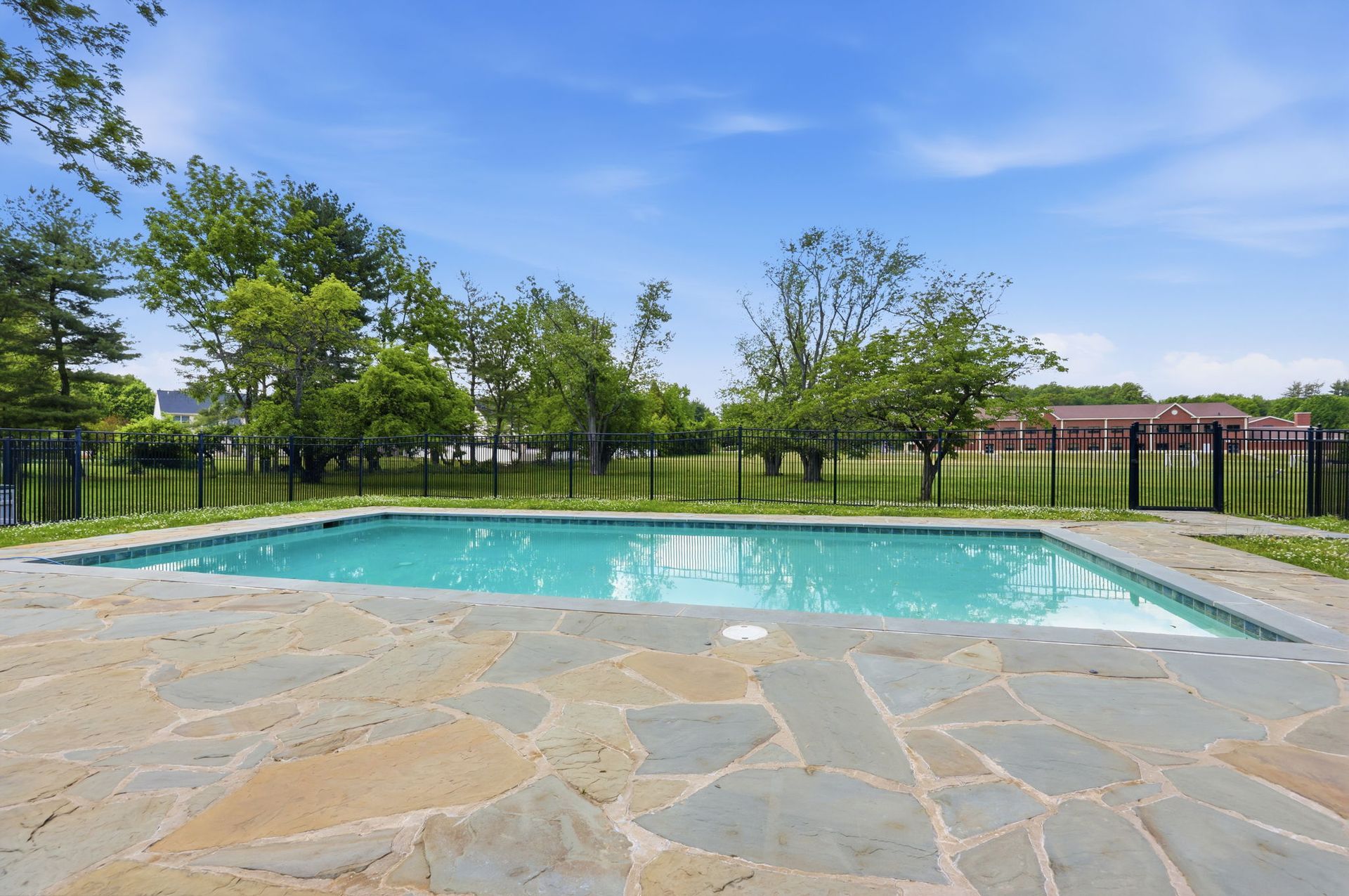 Empty outdoor swimming pool beside a stone patio, with green trees and a fence under a blue sky.