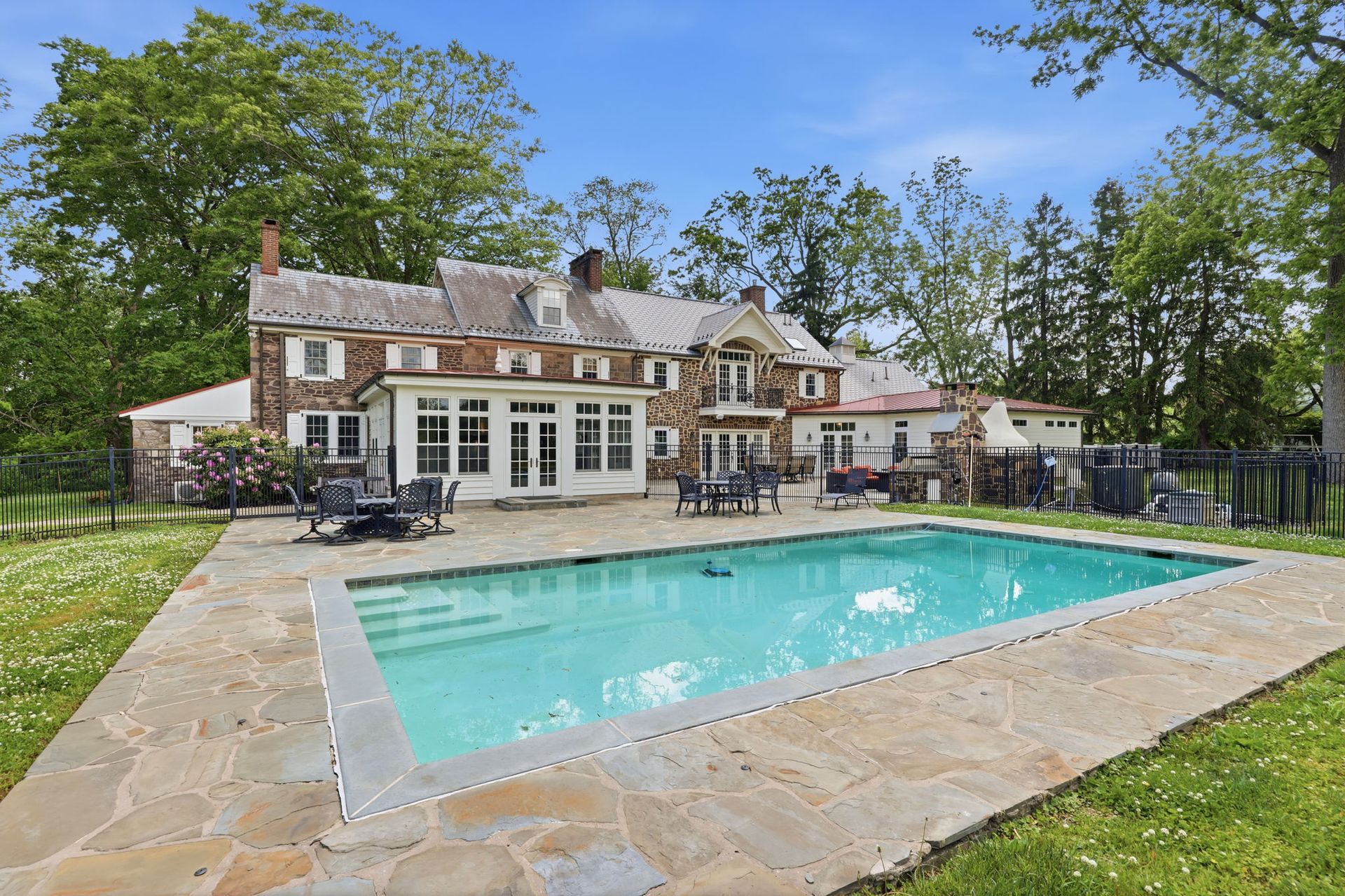 Backyard pool beside a stone patio and large house under a blue sky