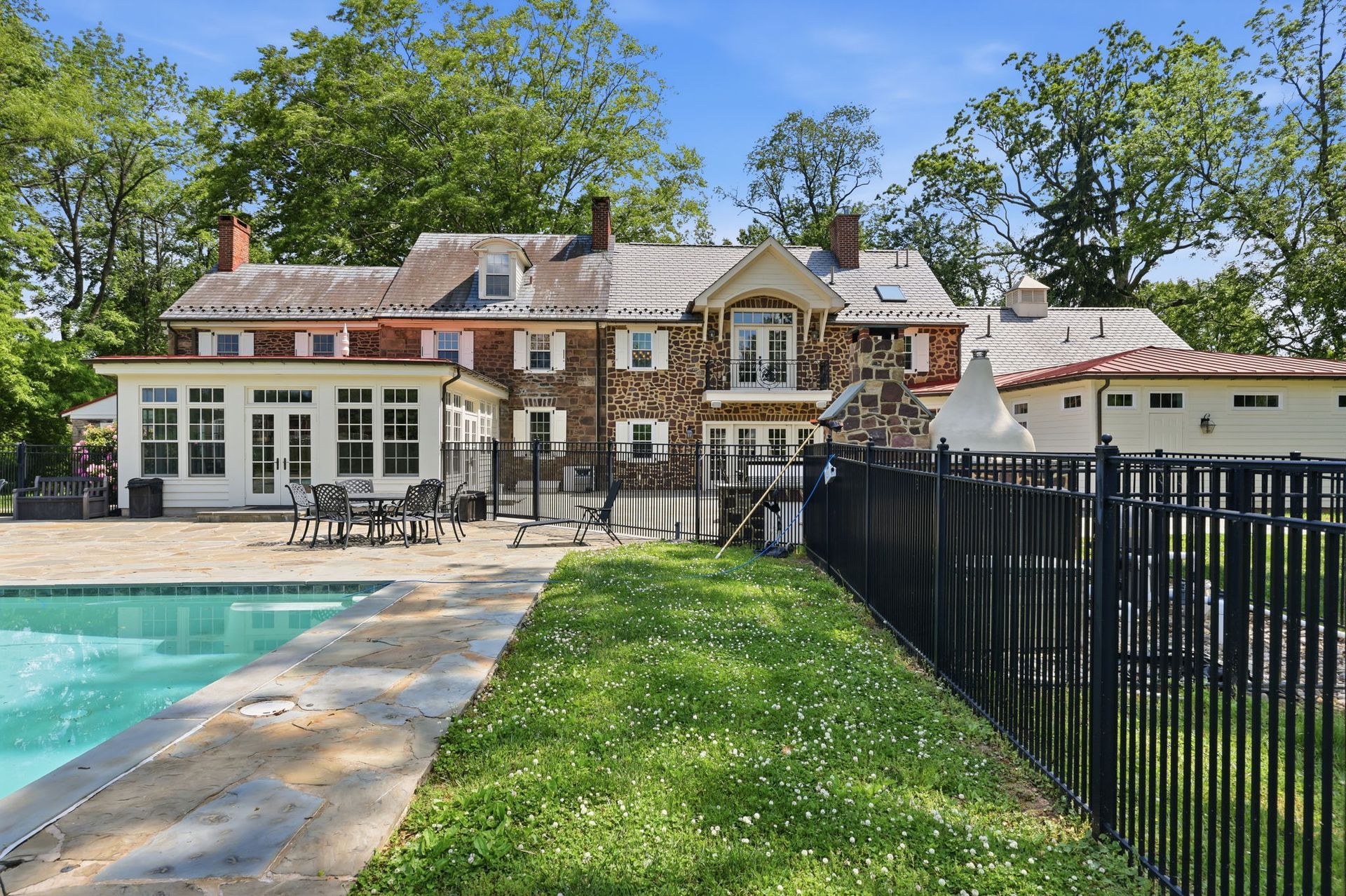 Outdoor pool beside a stone manor with patio seating, lawn, and black fence under a clear blue sky