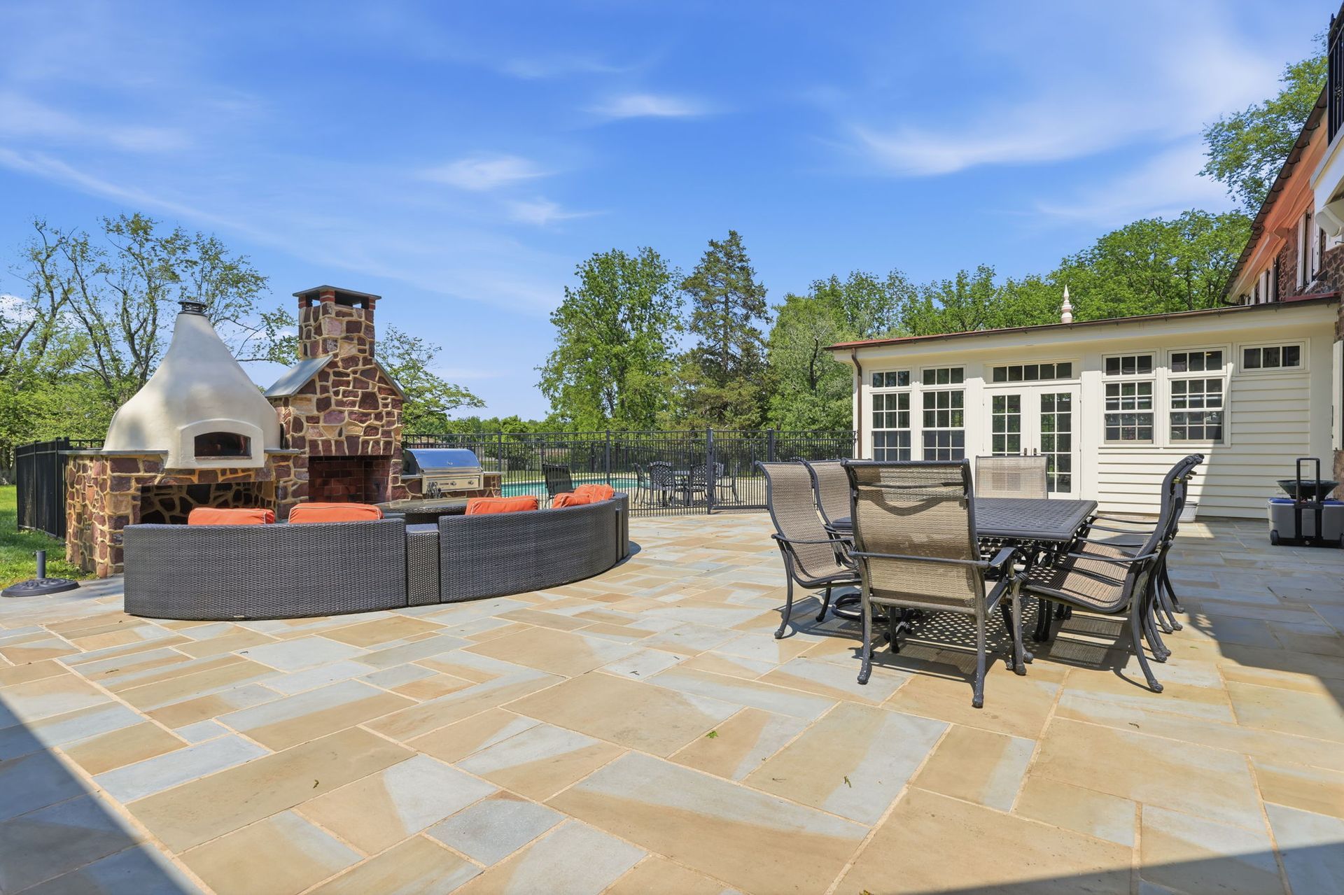 Patio with outdoor dining table, stone fireplace, and brick grill beside a white building on a sunny day