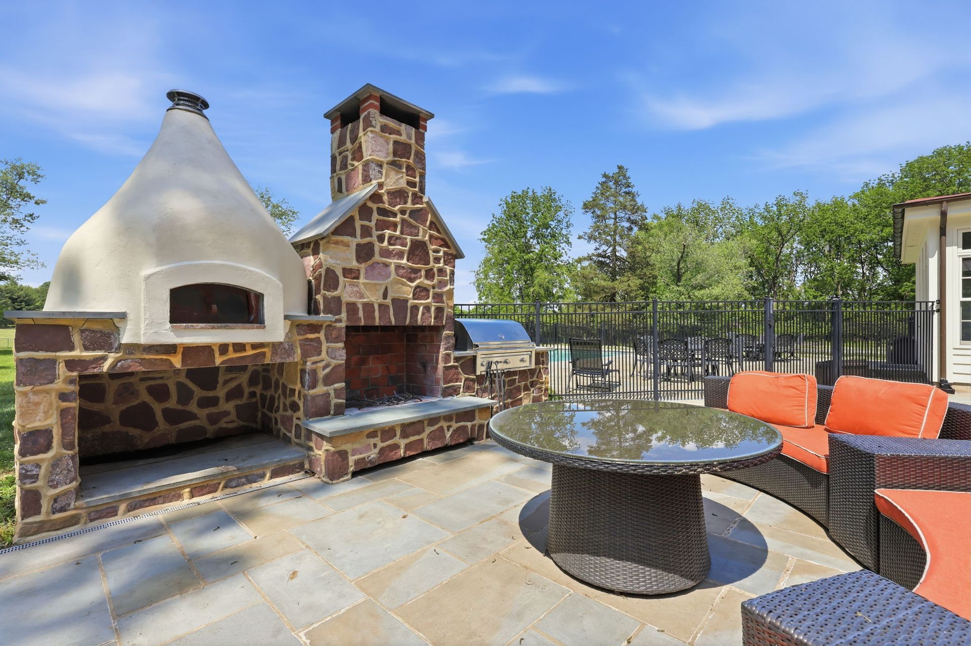Outdoor patio with stone pizza oven, brick fireplace, glass table, and orange lounge chairs under a blue sky