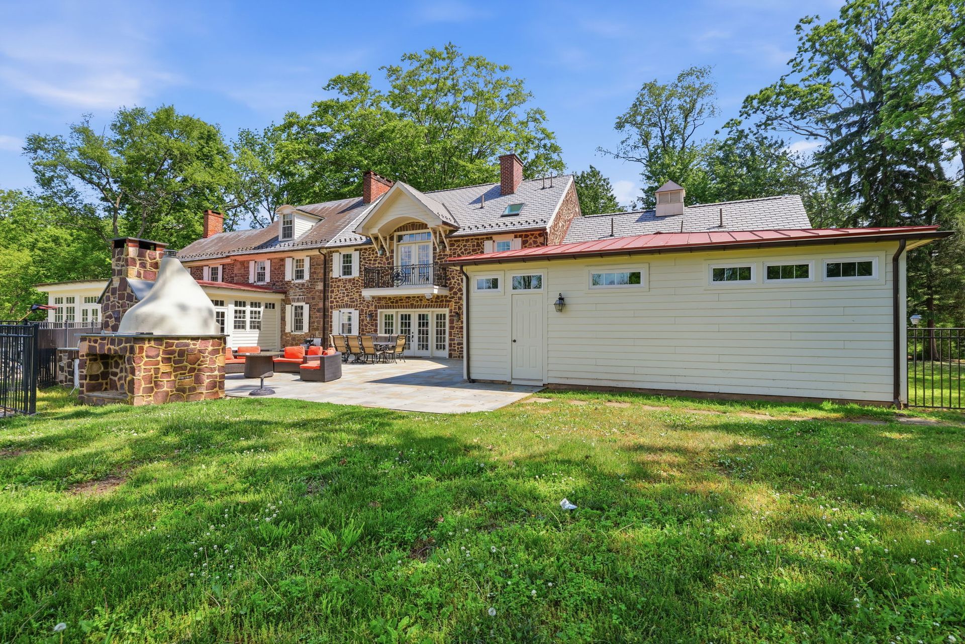 Stone house with a white garage, brick driveway, and green lawn under trees on a sunny day