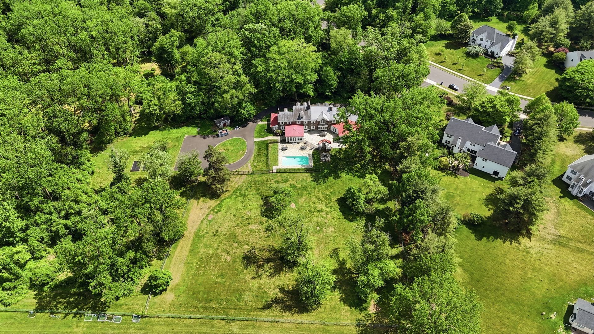 Aerial view of a wooded property with a house, swimming pool, and open grassy lawn